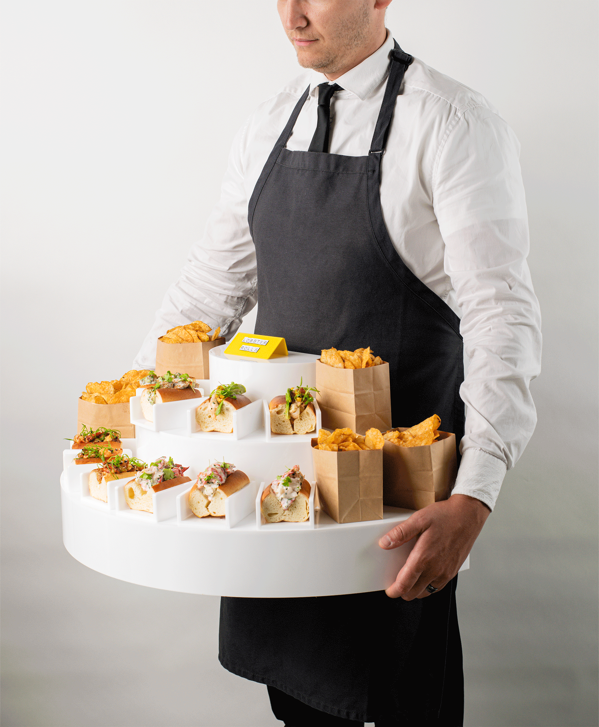 Man wearing a white shirt, black apron, and black tie holding a large round tray with various sandwiches and snack bags.