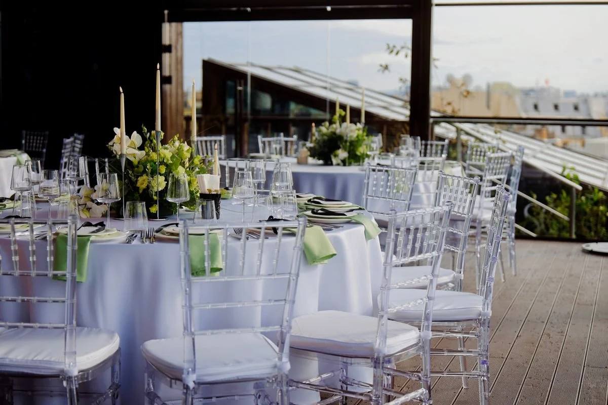A bunch of clear chairs surrounding tables adorned with a white tablecloth on a roof