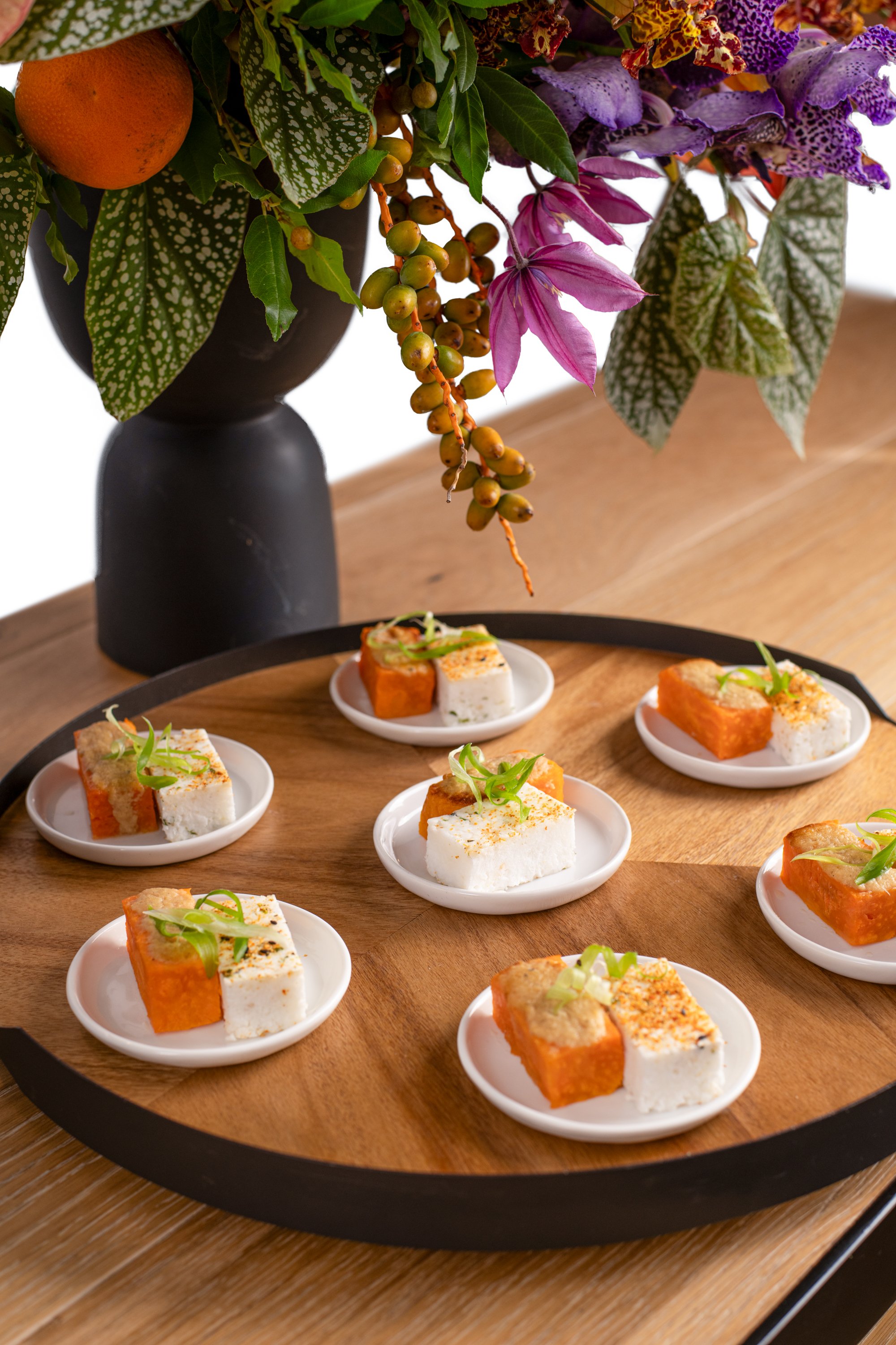 Small plates of assorted savory snacks on a wooden platter, with a large floral arrangement in a black vase in the background.