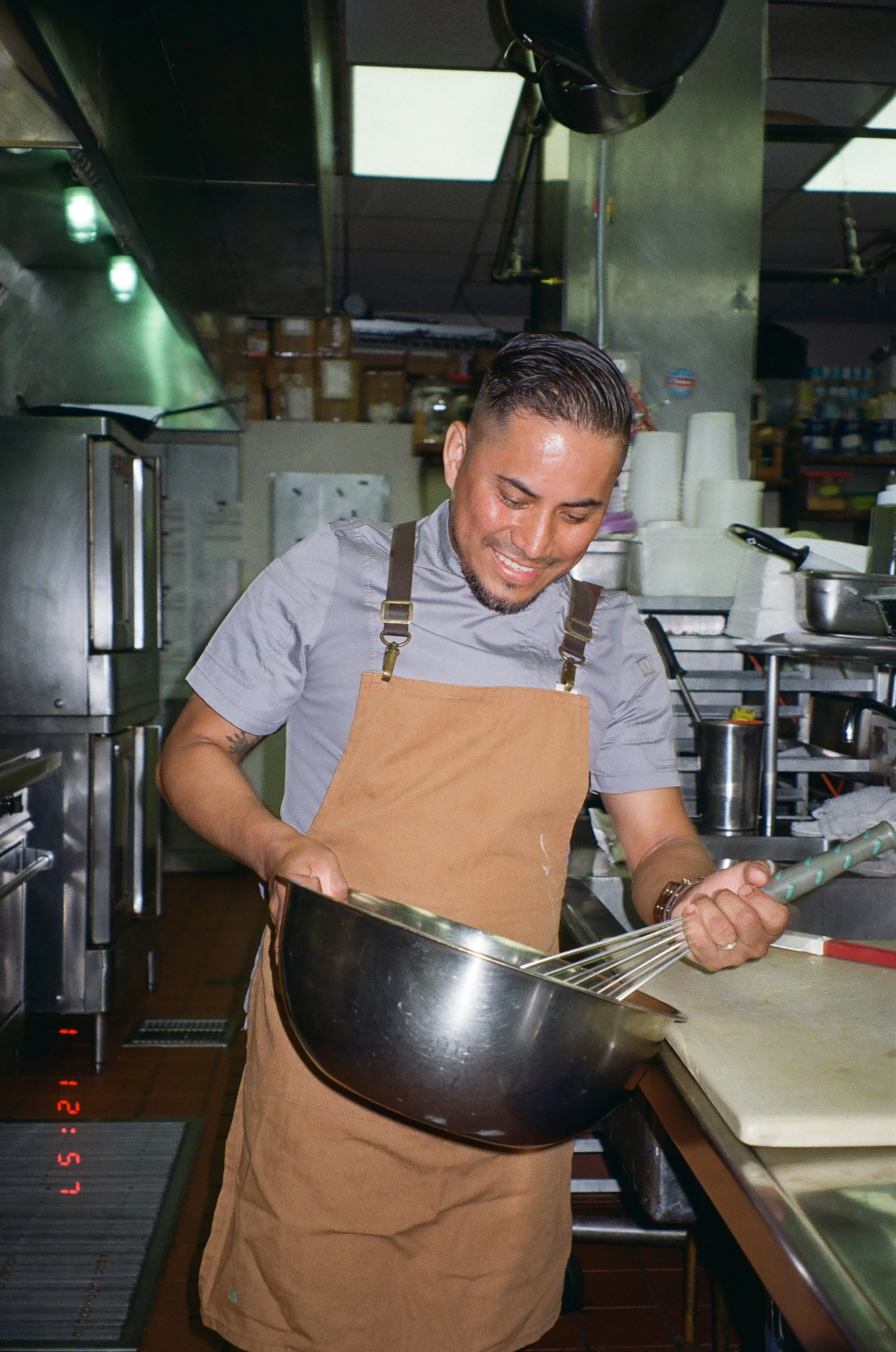 A man with a apron, smiling and whisking ingredients in a metal bowl in a professional kitchen.