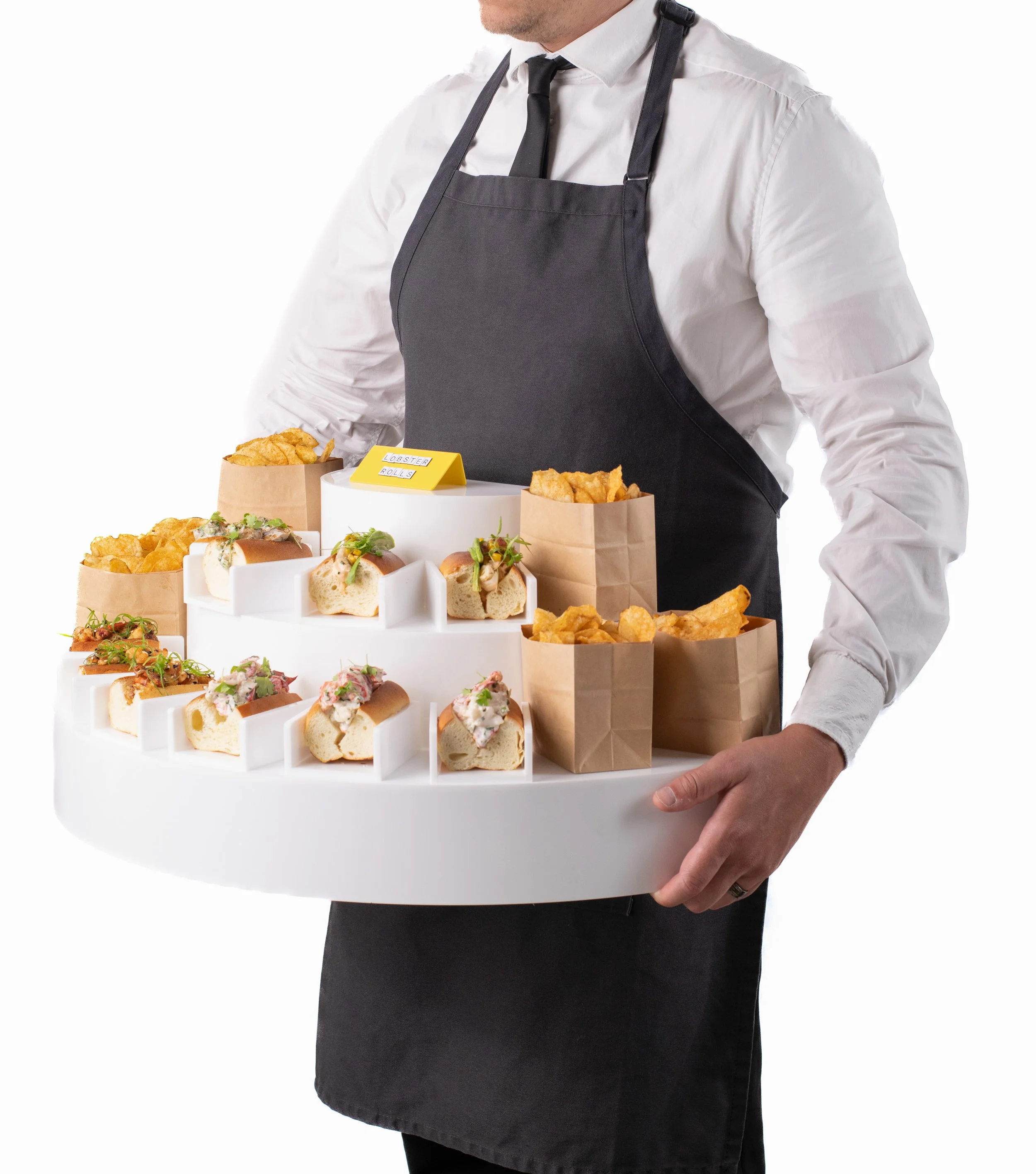 A person wearing a white shirt, black tie, and black apron holding a large round white tray with various mini sandwiches and brown paper bags of potato chips.