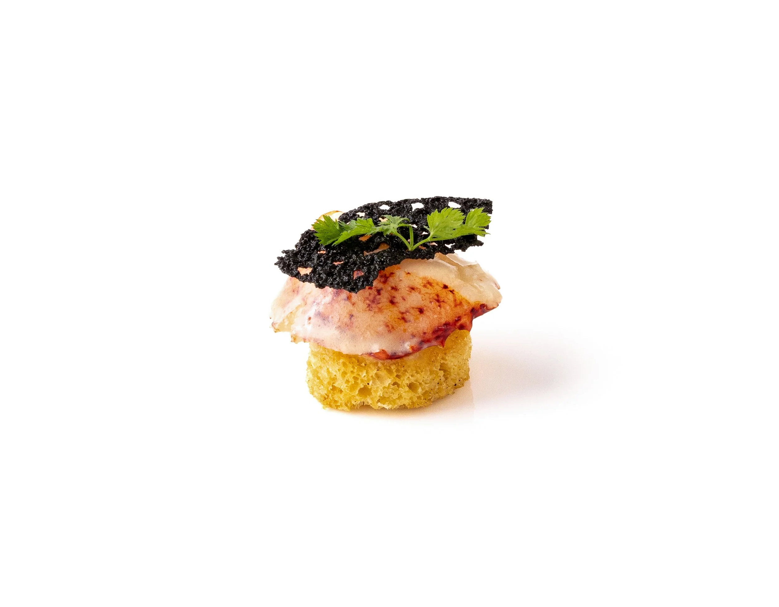 An artistic appetizer with a small piece of bread, topped with a slice of pink seafood, a green leafy garnish, and a black crispy element on top, all arranged on a white background.