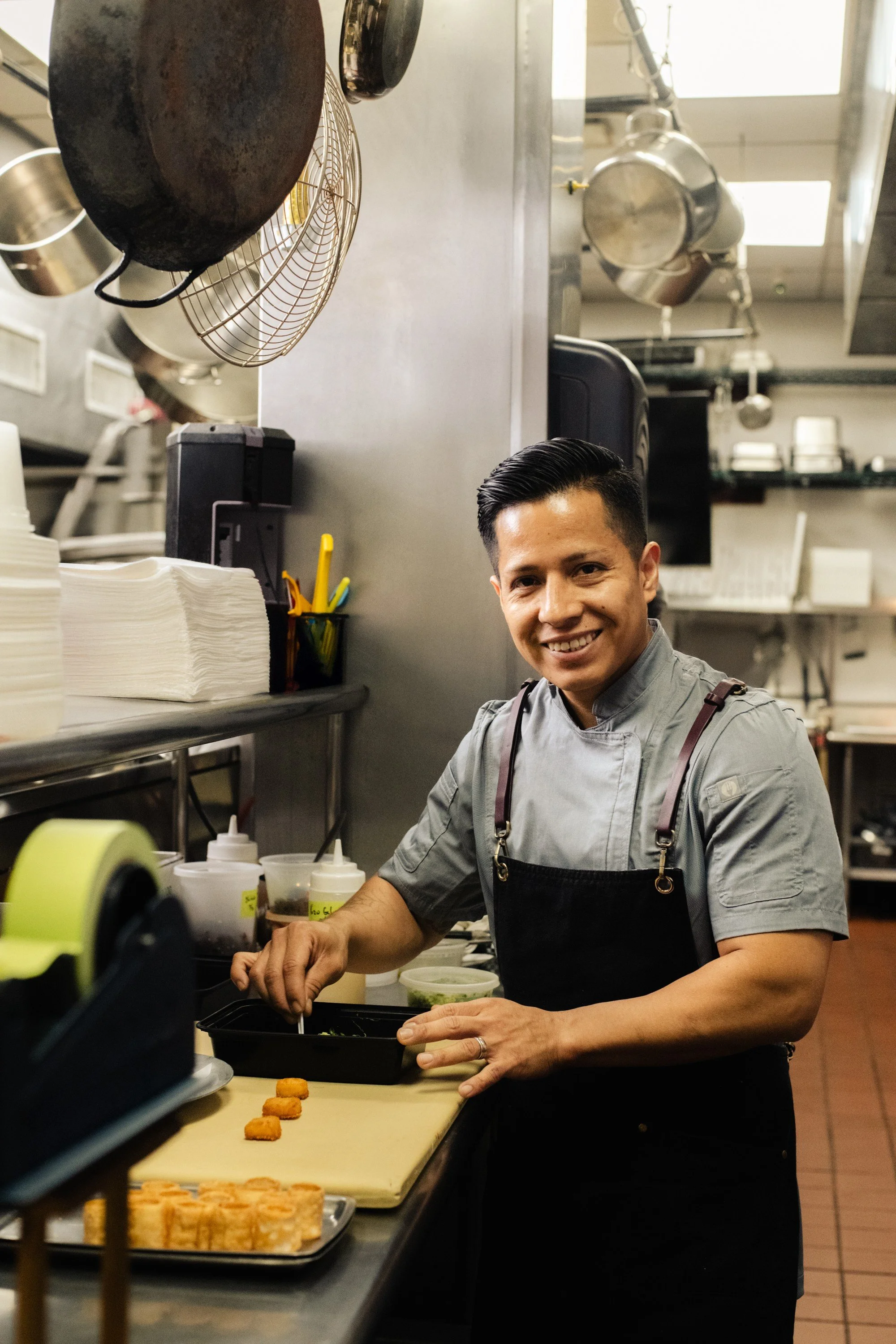 A smiling chef preparing food in a commercial kitchen, with various kitchen utensils, containers, and ingredients surrounding him.