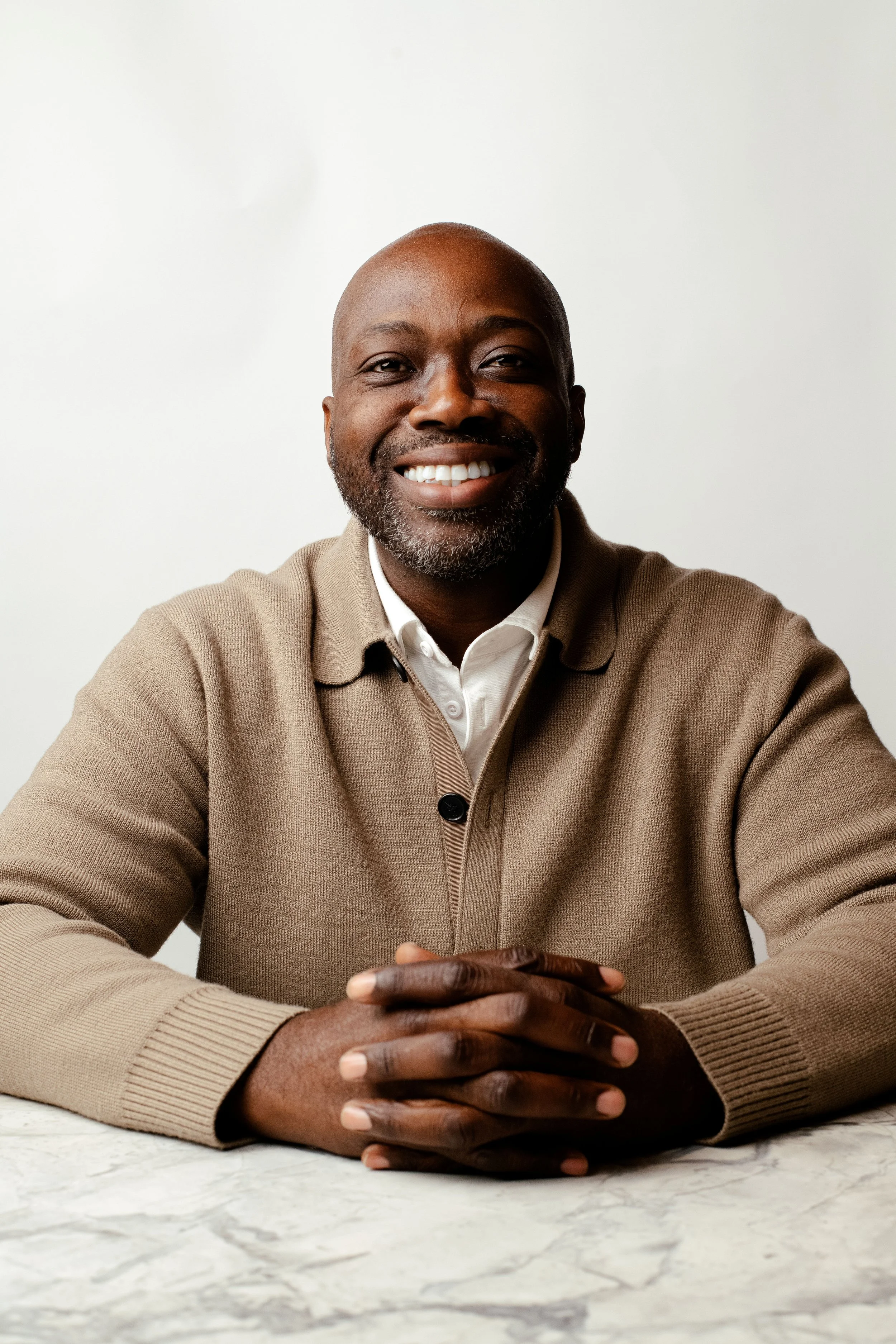 Smiling man wearing a beige cardigan and white shirt, sitting at a marble table against a plain white background.