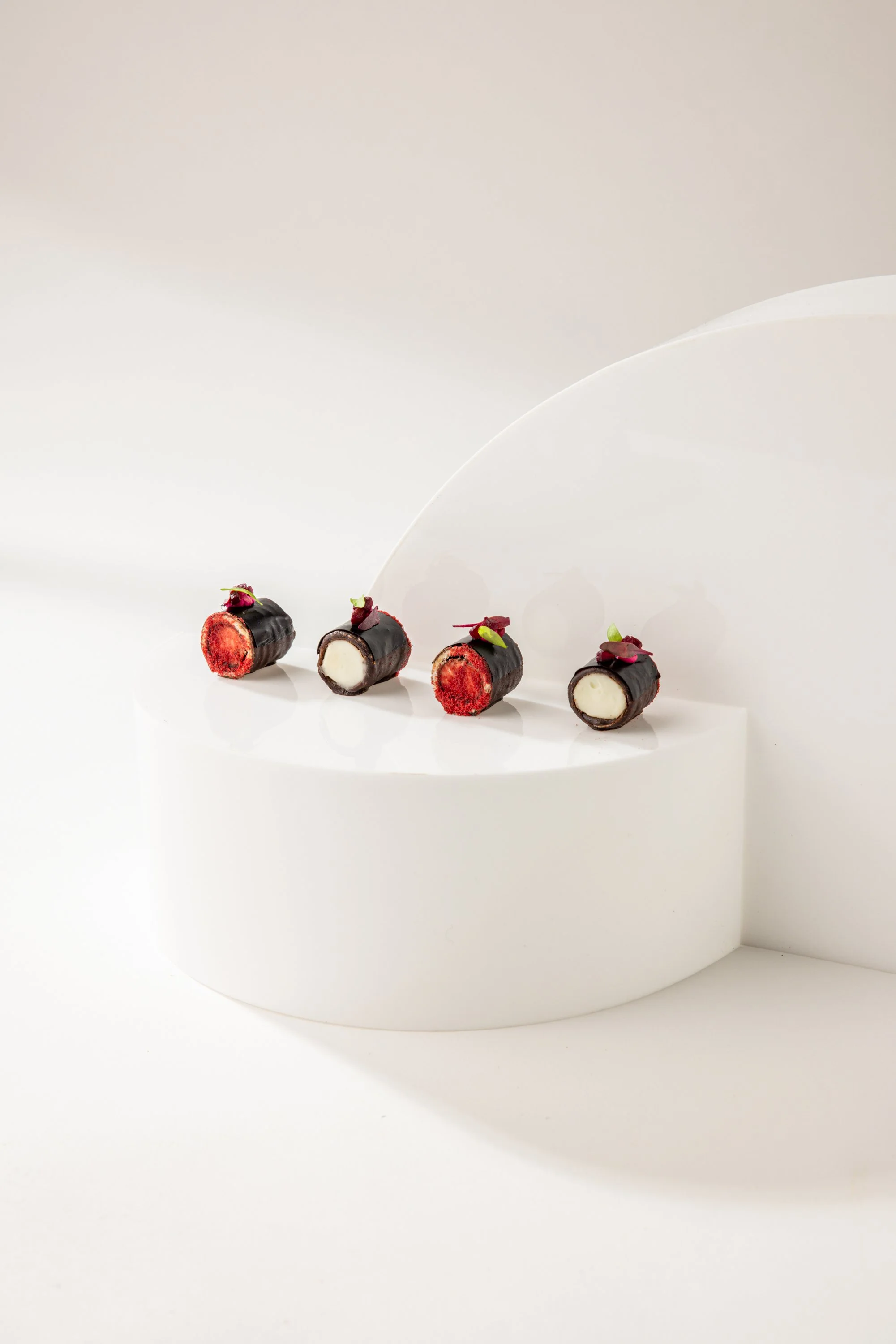 Four small cylindrical desserts with dark chocolate coating, filled with red and white fillings, and garnished with small green and purple leaves, arranged on a white curved display against a white background.