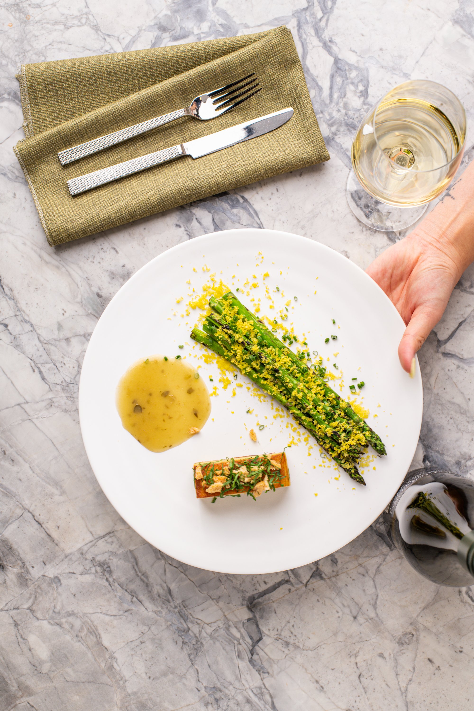 A white plate with grilled asparagus topped with lemon zest, a small rectangular piece of salmon garnished with herbs, a yellowish sauce, and a glass of white wine on a marble table.