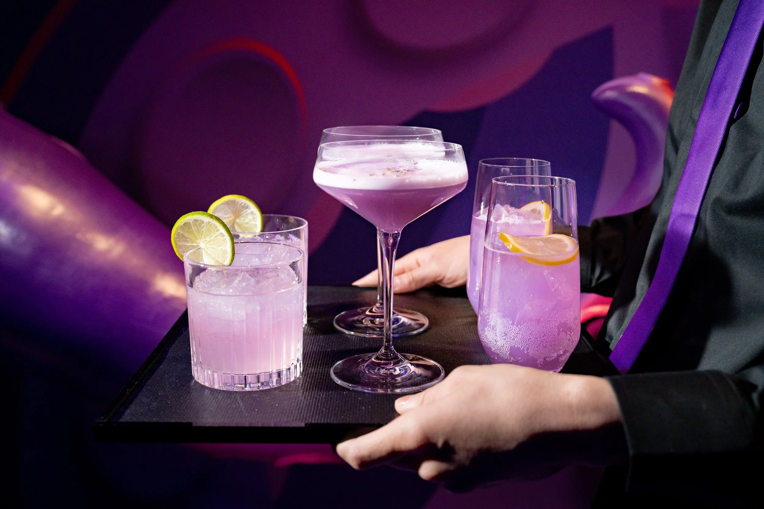 A server carrying a tray with four glasses of pink and purple cocktails, garnished with lemon and lime slices, in a dimly lit lounge or bar setting.
