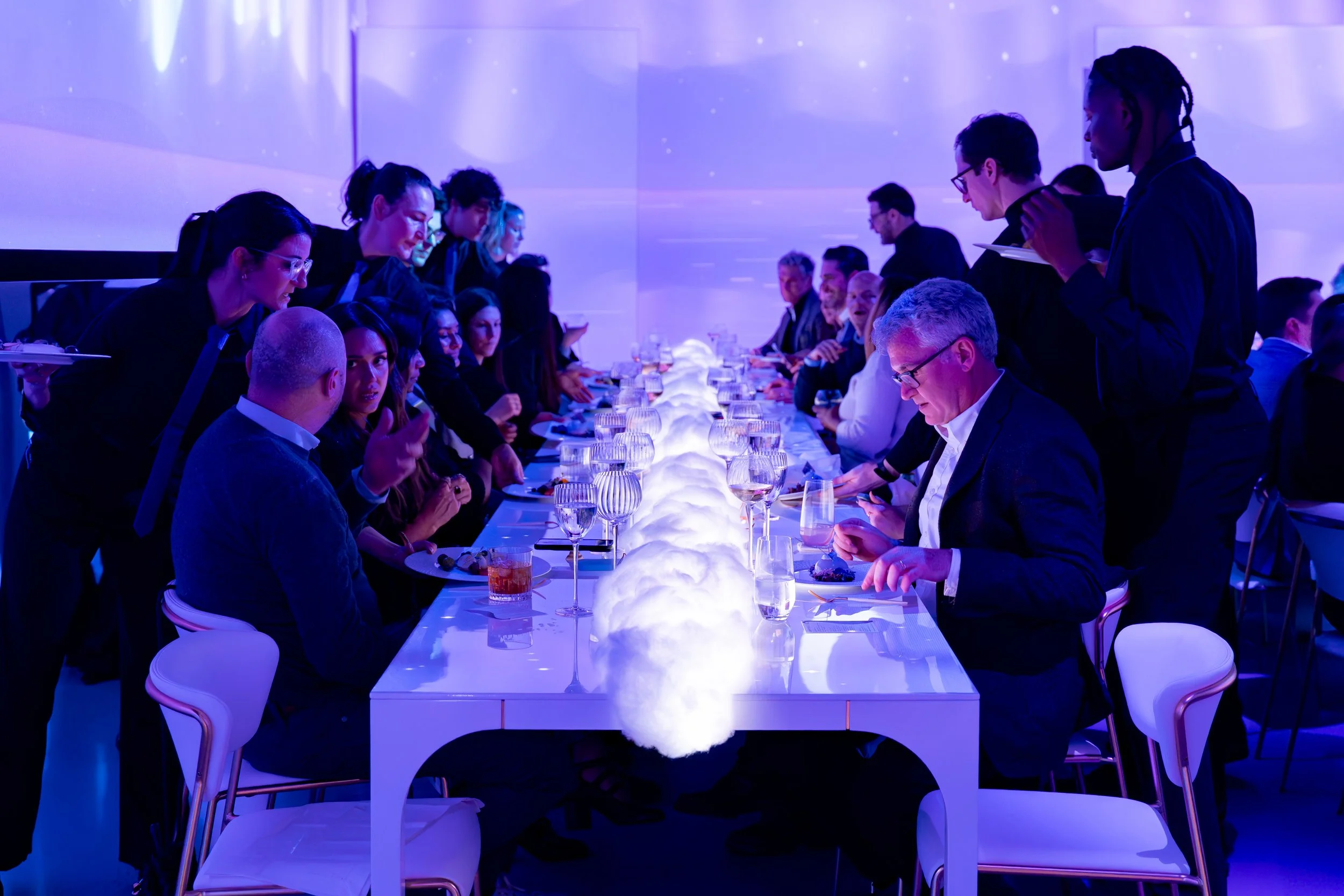People seated at a long dining table with glowing cloud-like decorations in the center, under blue-purple lighting, at a formal event or dinner party.