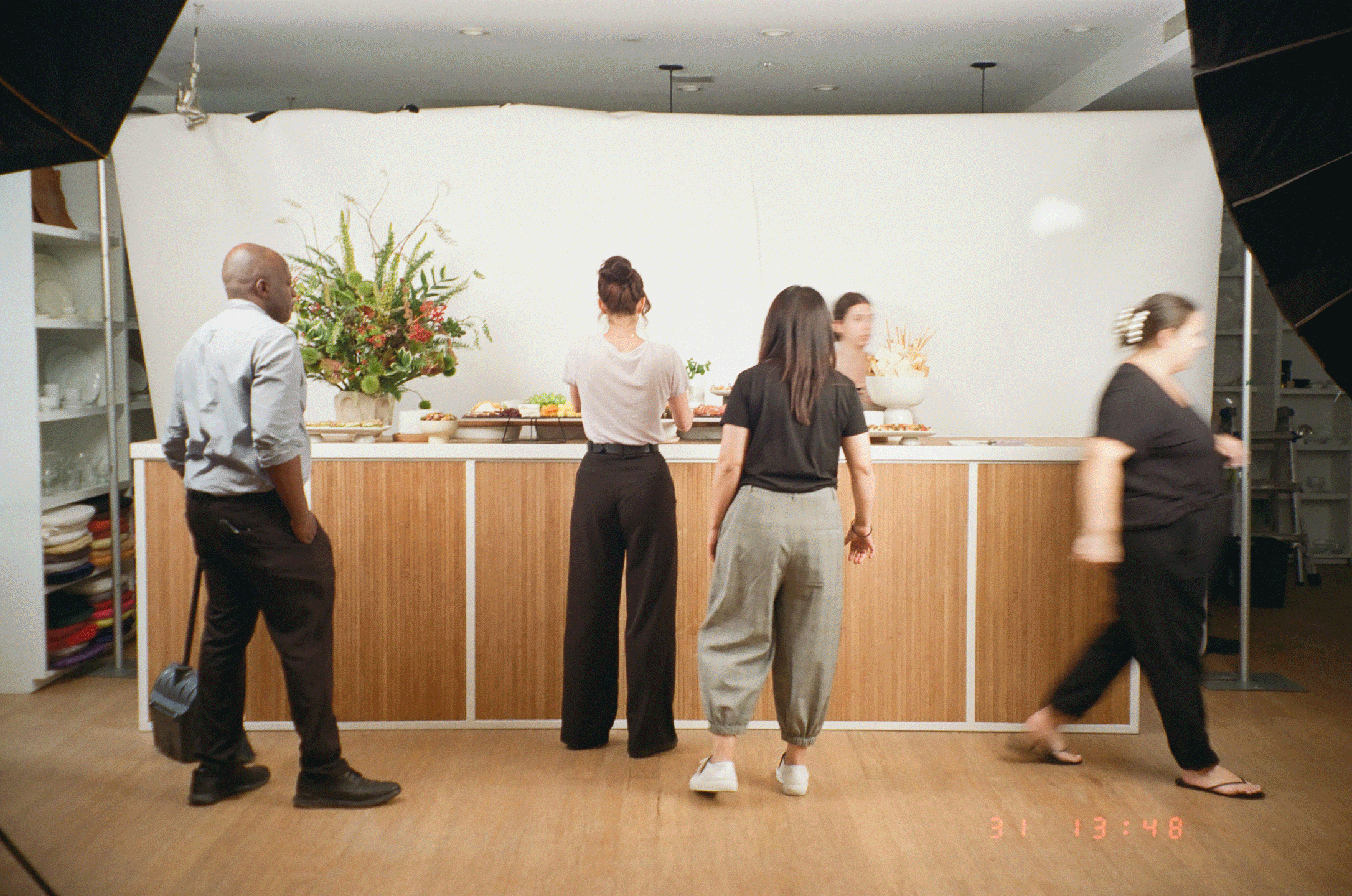 People standing in line at a buffet table with floral arrangement and food dishes, in a room with white walls and shelving on the side.