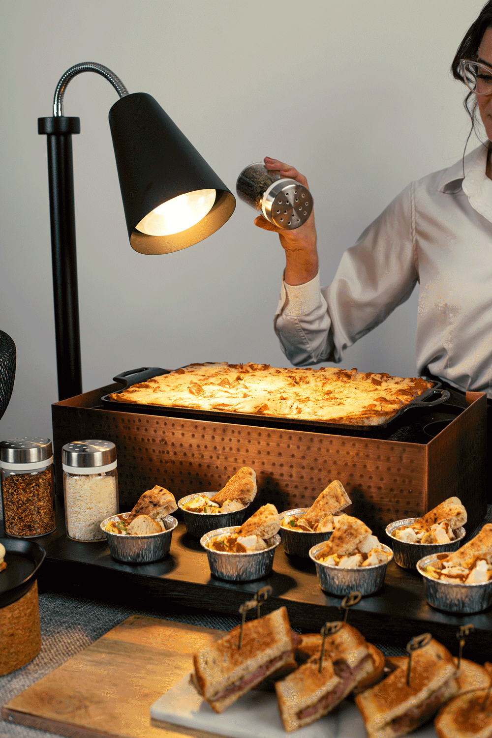 A person operating a food warmer with a tray of baked cheesy bread, surrounded by sandwiches and small bowls of dip or sauces.
