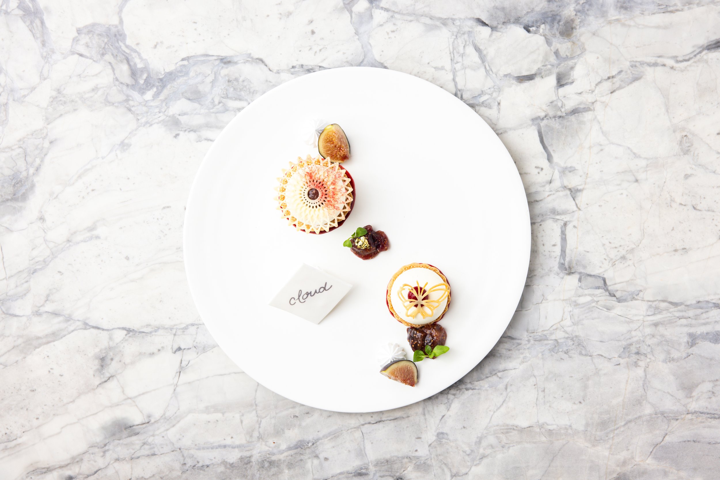 Elegant dessert plate with various pastries, garnished with figs, herbs, and decorative elements, on a white marble surface.