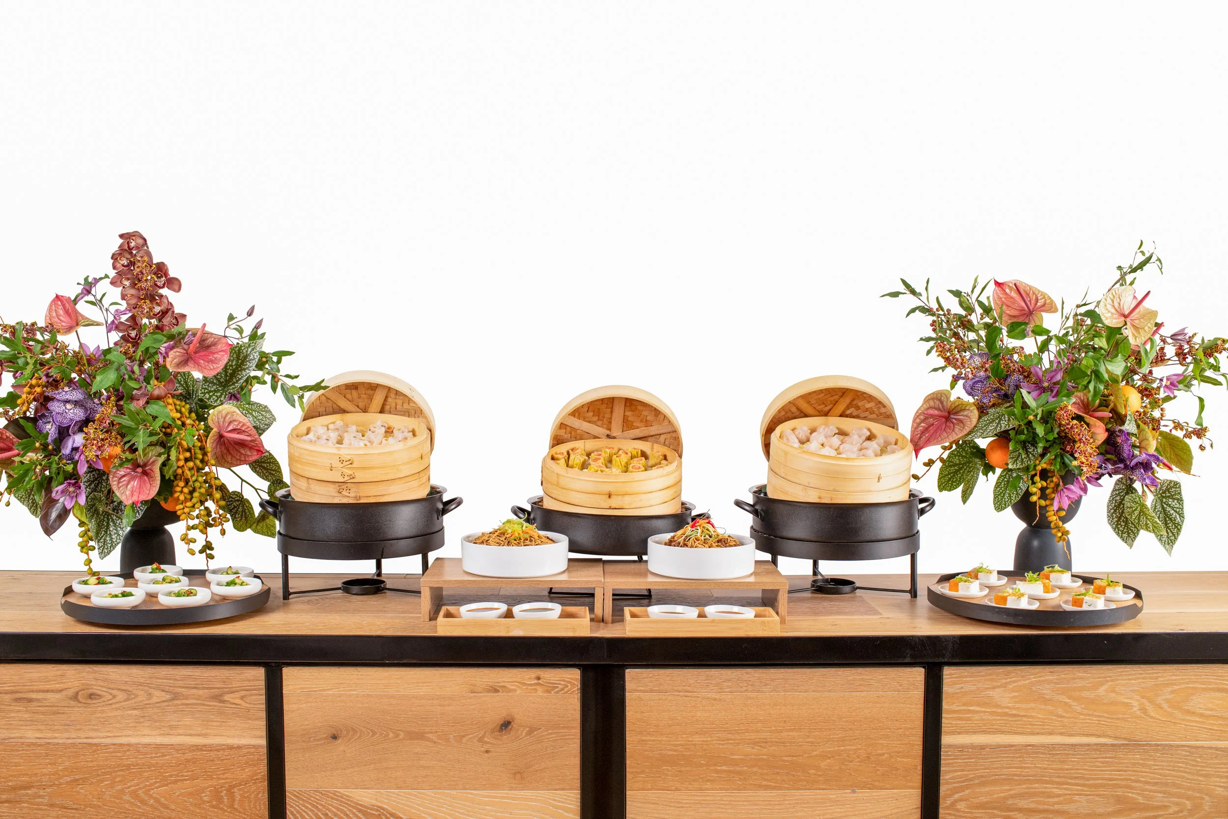 Arrangement of Asian cuisine dishes with dim sum and noodle bowls, flanked by colorful floral arrangements on a wooden table.