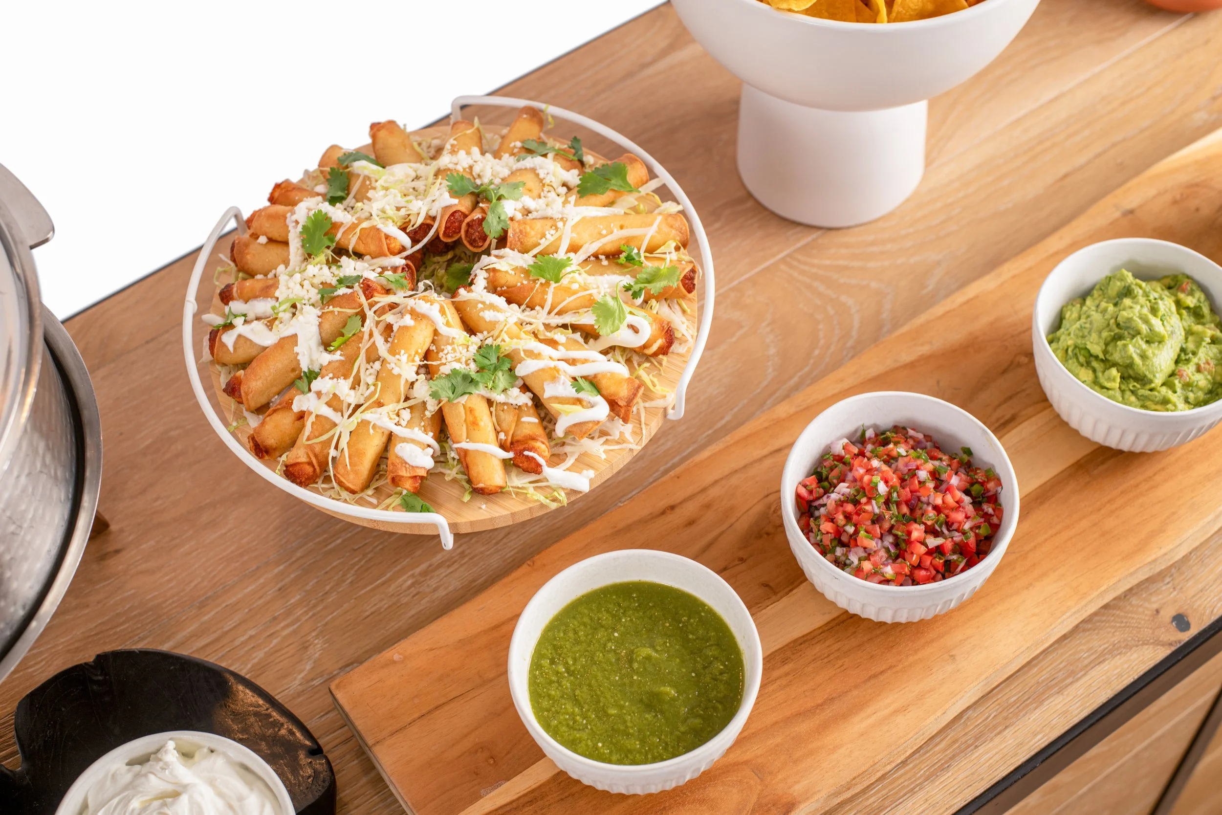 Plate of taquitos garnished with cilantro and cheese, served with bowls of salsa verde, pico de gallo, and guacamole on a wooden serving board.