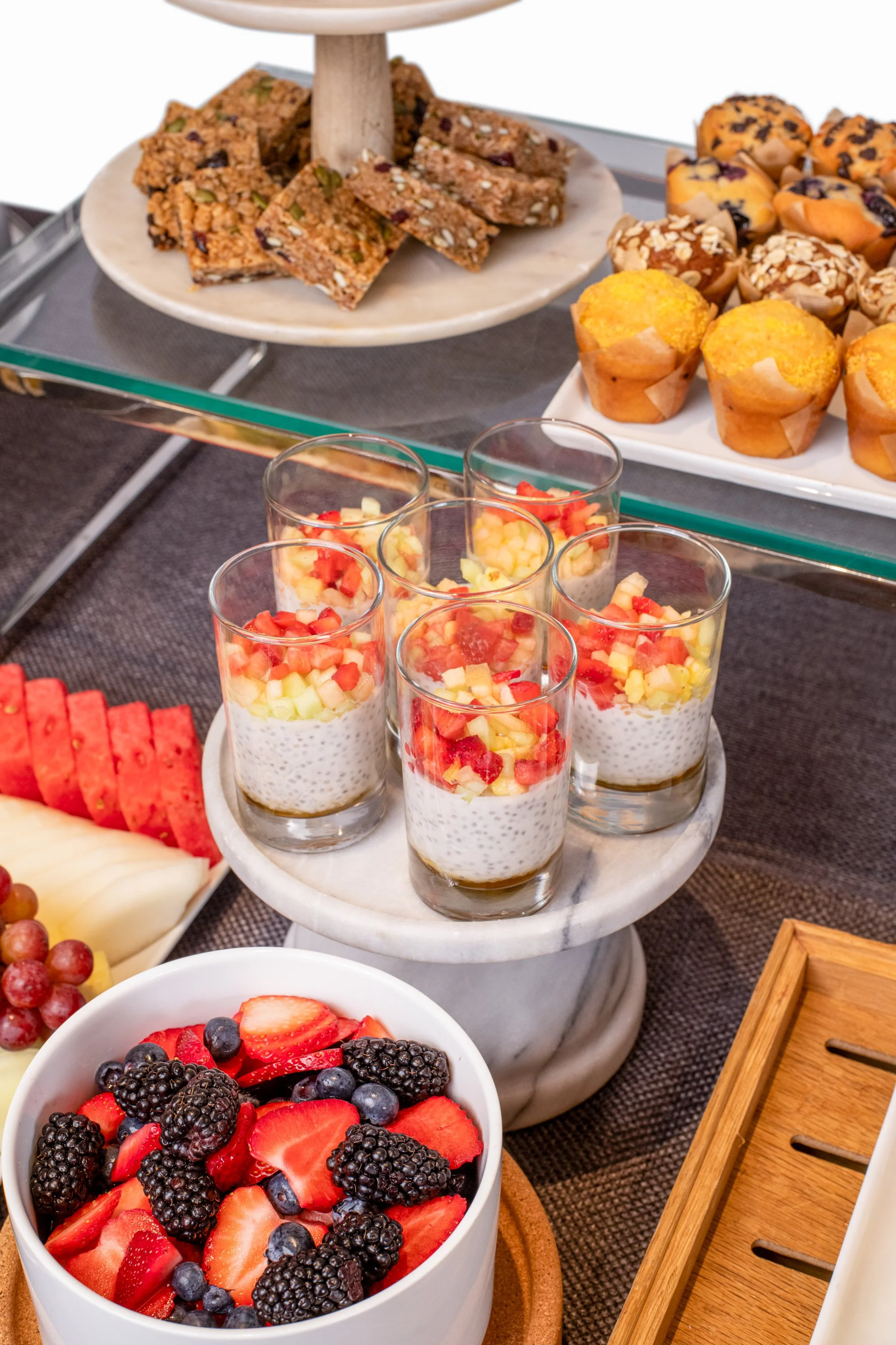 Assorted desserts including chia seed pudding topped with fruit in glasses, watermelon, honeydew, and cantaloupe slices on a tray, chocolate chip muffins, fruit bars on a tiered stand, and a bowl of mixed berries.