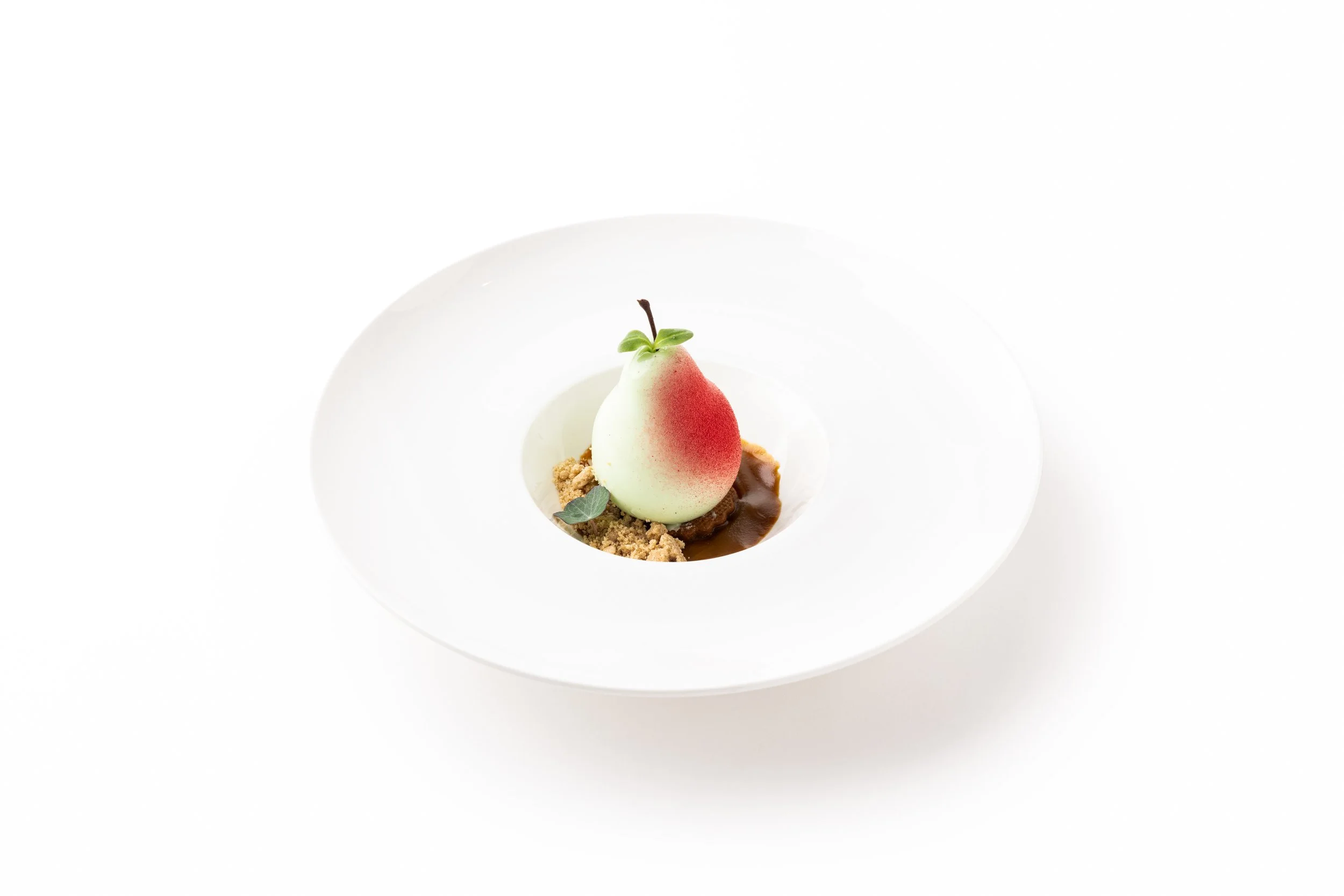 Elegant dessert featuring a pear-shaped element with a red and white gradient, garnished with a small green leaf, served on a white plate with crumb and sauce accents.