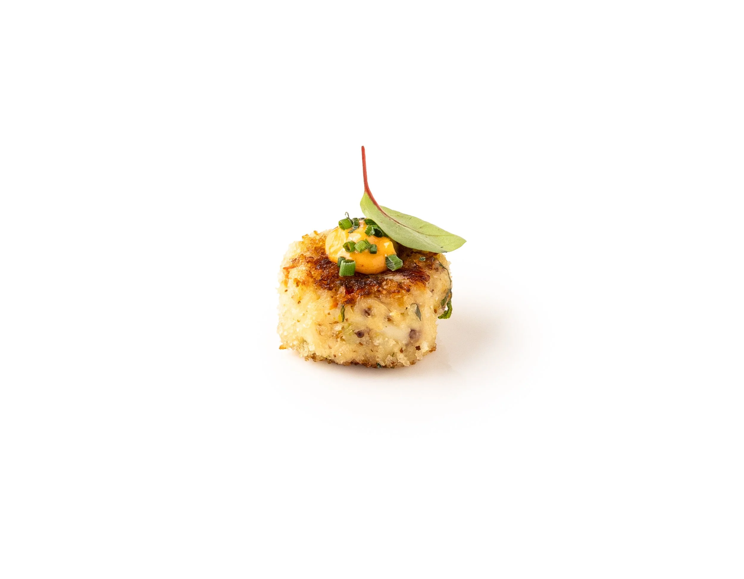 A small, square piece of breaded and fried food garnished with a leaf, chopped green herbs, and a dollop of yellow sauce on top, white background.