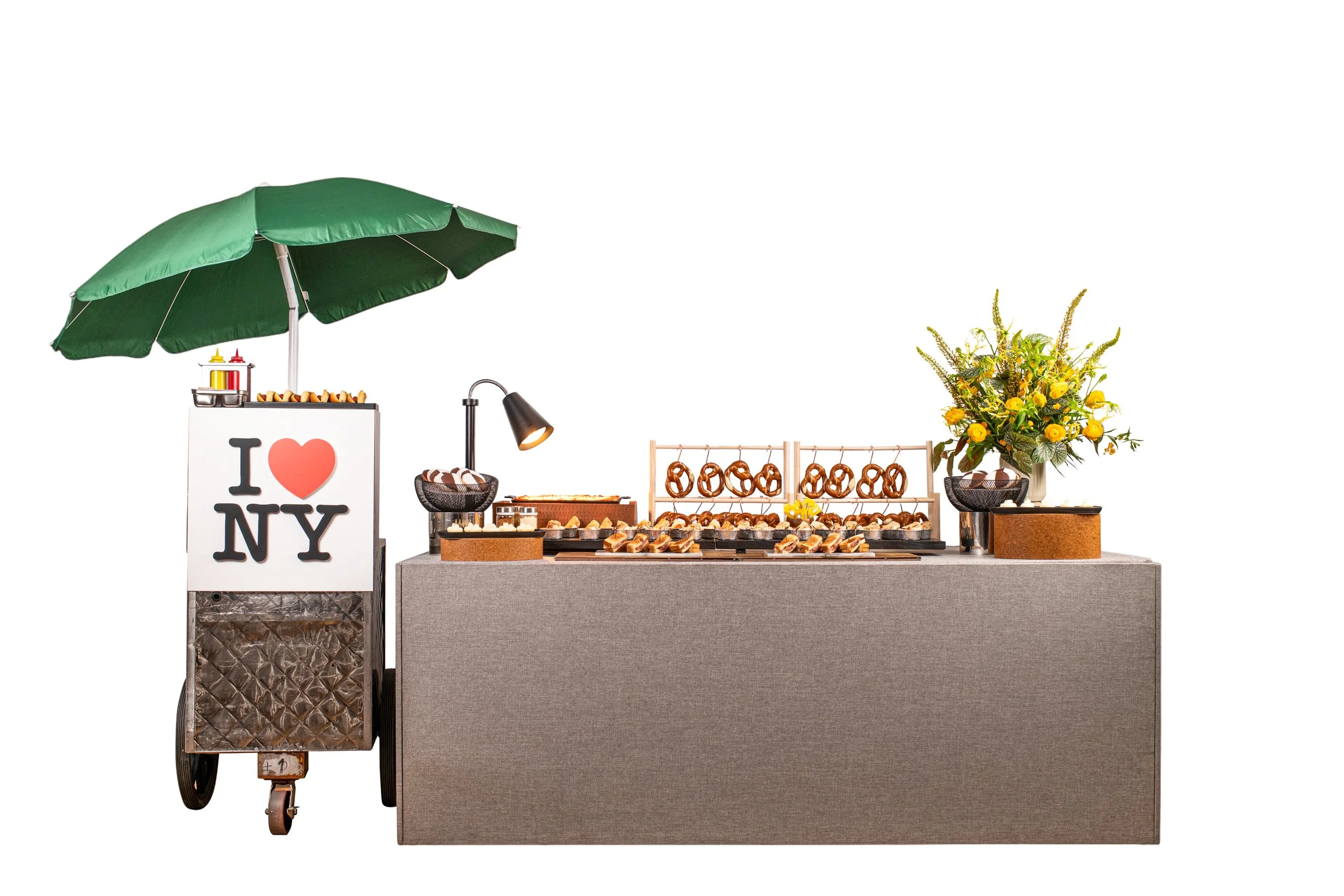 A street food cart with a green umbrella featuring a sign that says 'I love NY' in black and red, and an assortment of baked goods like pretzels, sandwiches, and other snacks displayed on the cart. There is also a large floral arrangement with yellow