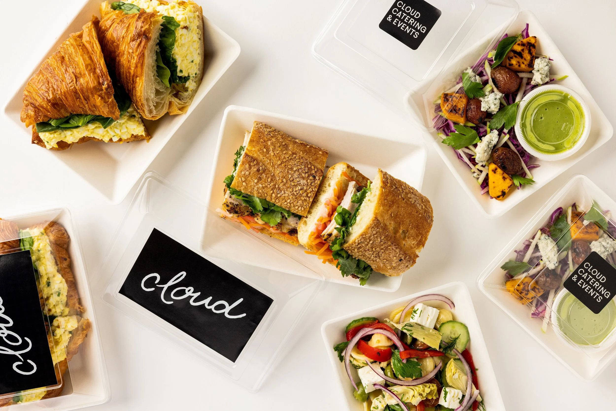 Assorted sandwiches, salads, and snacks in white plastic containers on a white surface, with some labeled 'cloud' and 'cloud catering & events'.