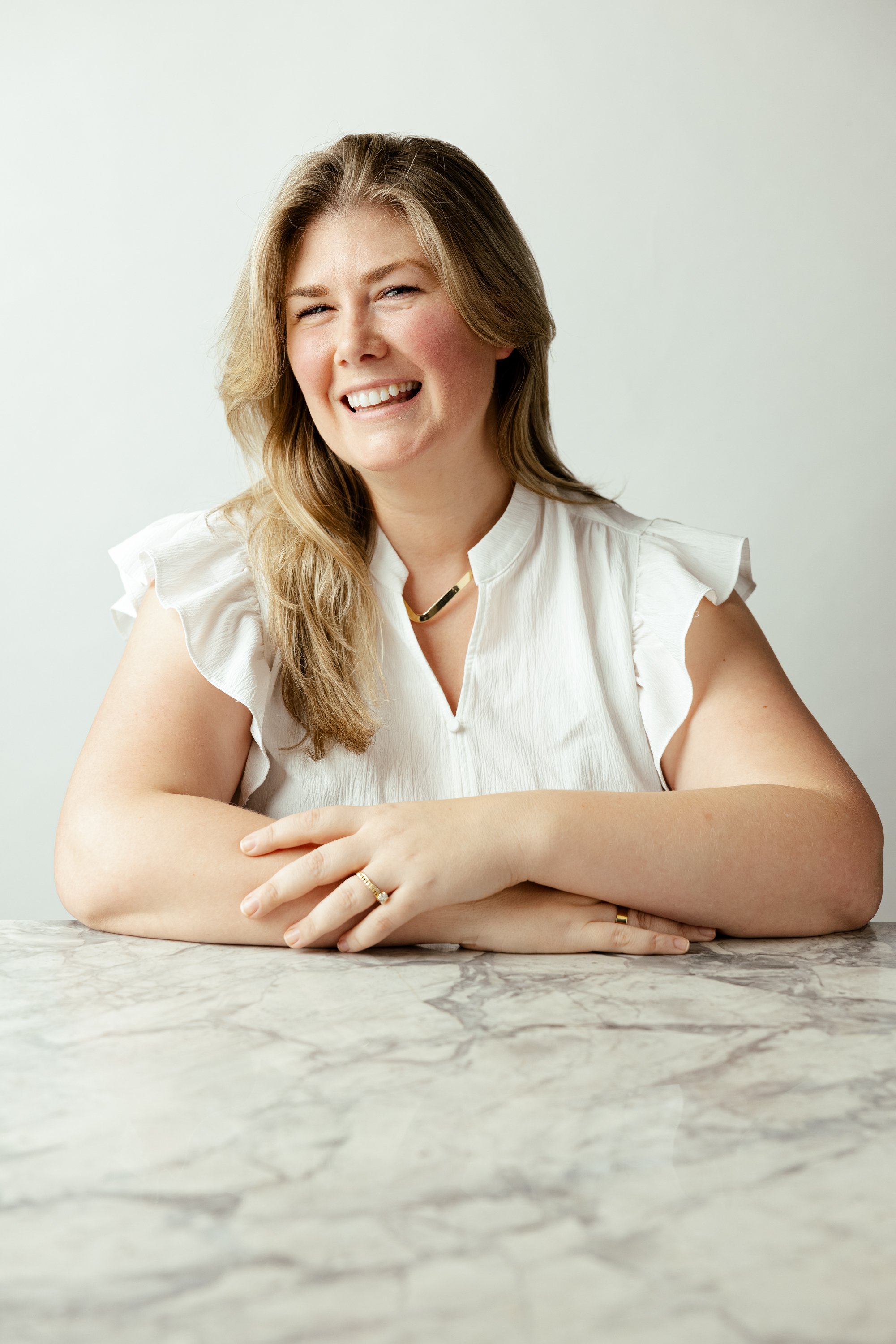 A woman with long, wavy blond hair smiling and sitting at a marble table in a white top with ruffled sleeves.