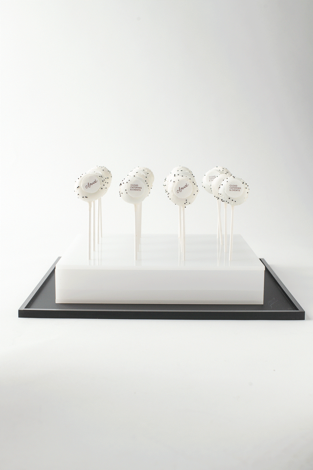 Four white cake pops with black sprinkles and a small round label reading 'Cloud' or 'Cloud Catering & Events' on each, displayed on a white platform with a black border against a plain light background.