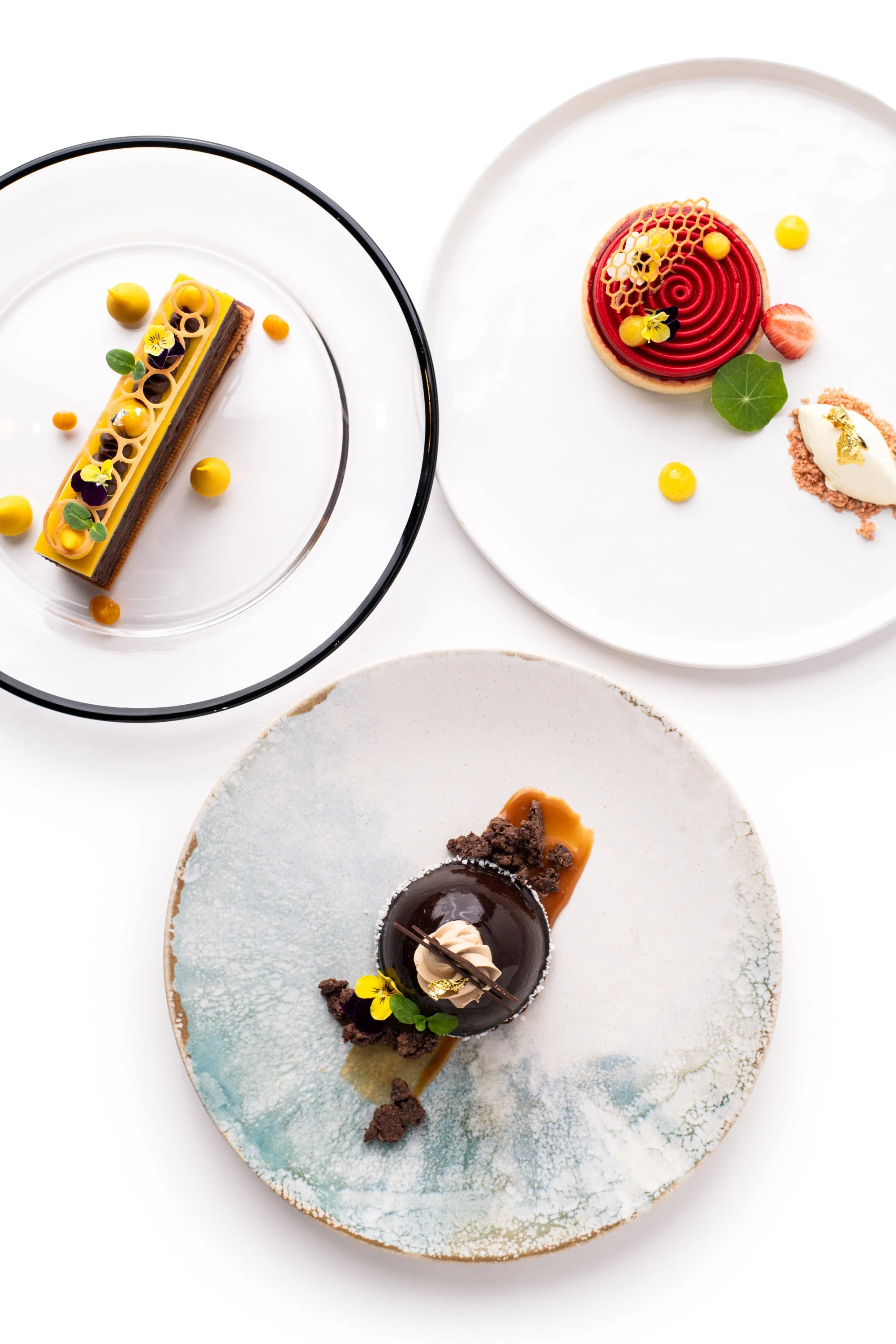 Three elegantly plated gourmet desserts on white plates, featuring various colorful toppings and garnishes.
