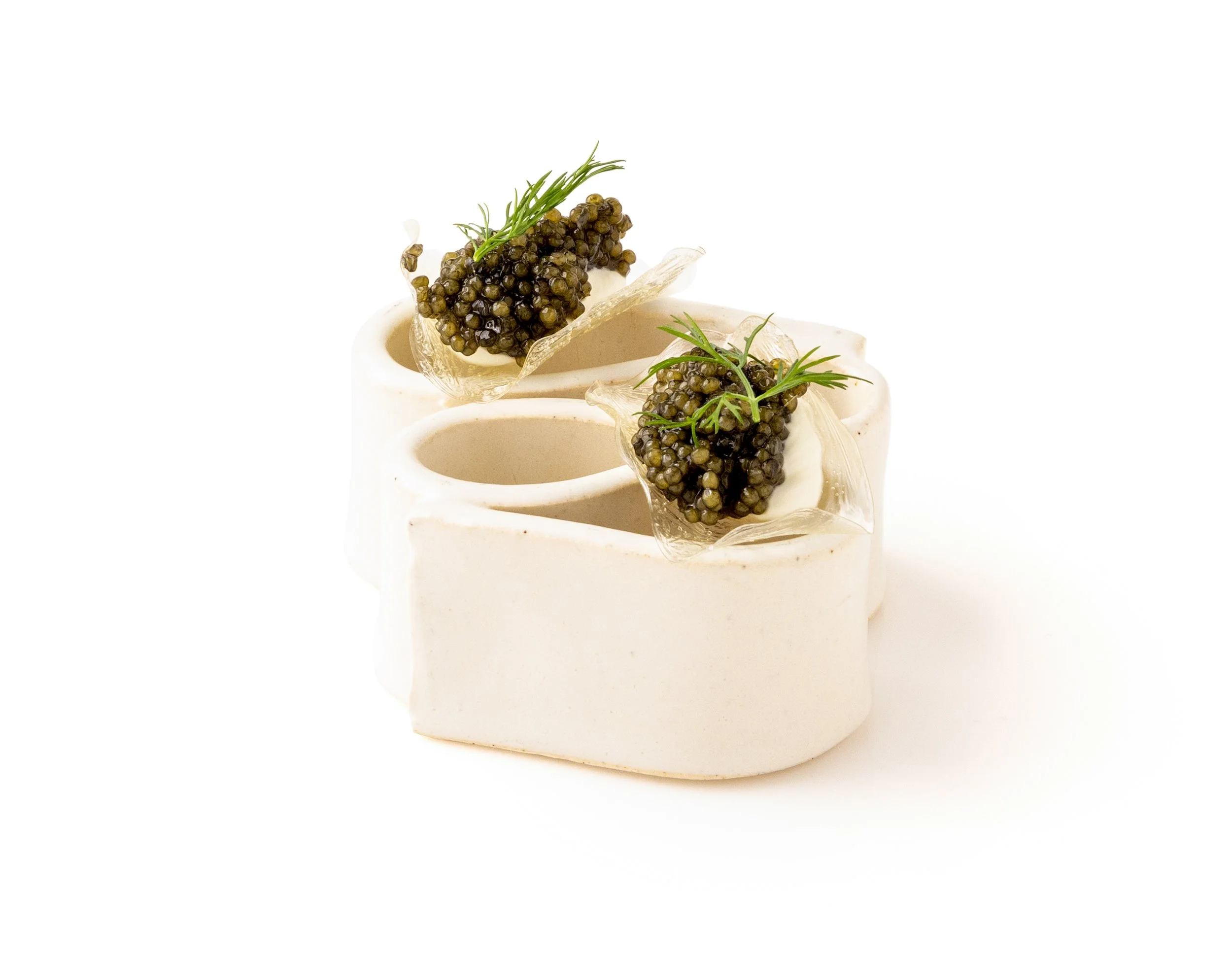 Two white ceramic containers each holding a spoon with black caviar and a sprig of green herbs, placed on a white background.