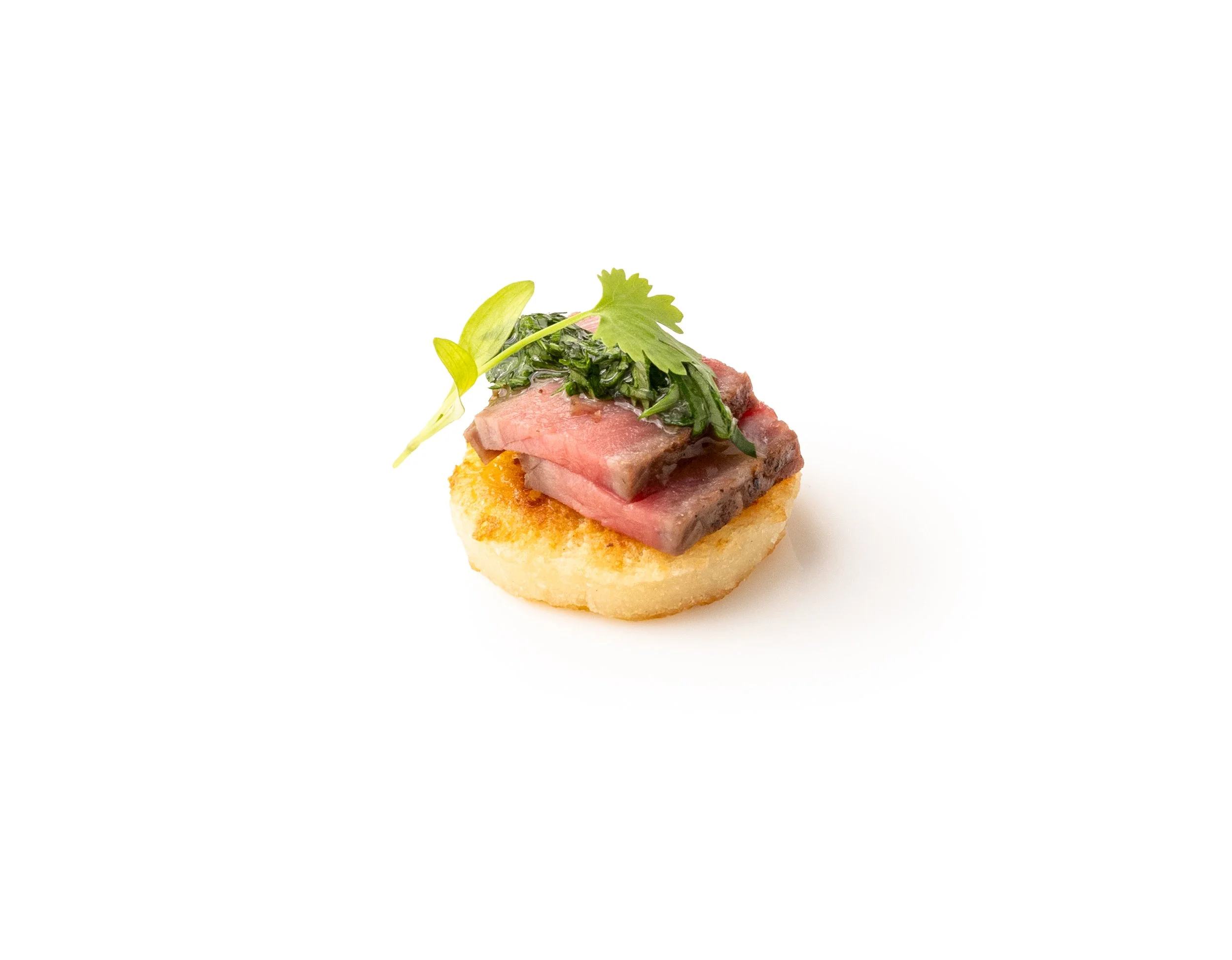 A small appetizer with a round, crispy base topped with cooked steak slices, fresh herbs, and microgreens, served on a white background.