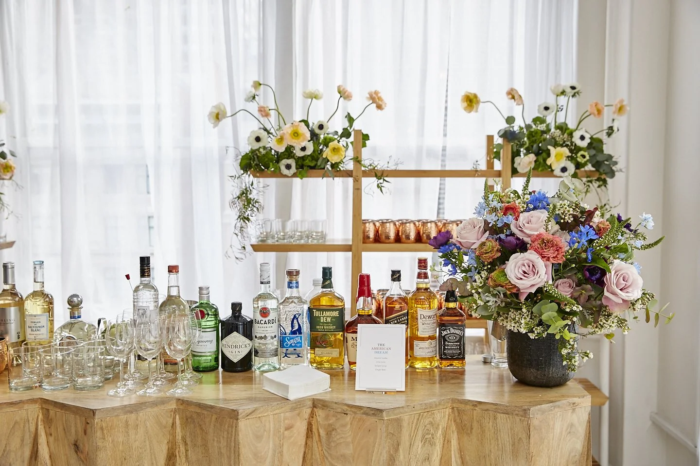 A bar setup with various liquor bottles, glassware, and floral arrangements on a wooden table in front of a window with sheer curtains.
