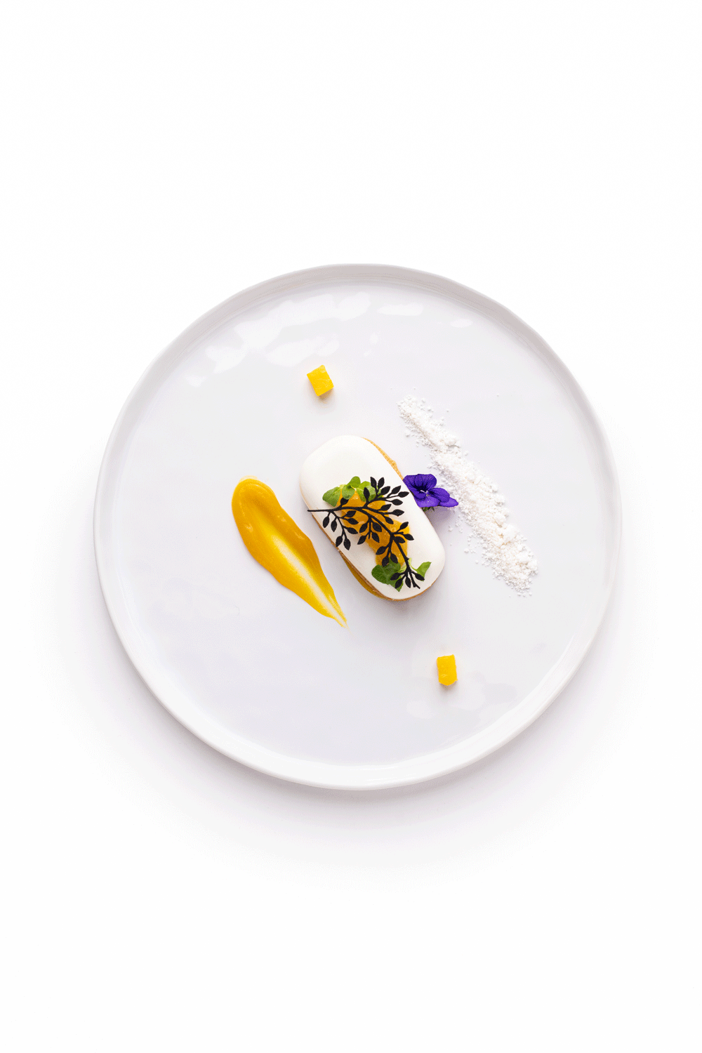 Elegant plated dessert with white mousse, yellow sauce, edible flowers, black decorative leaf, and small yellow garnish on white ceramic plate.