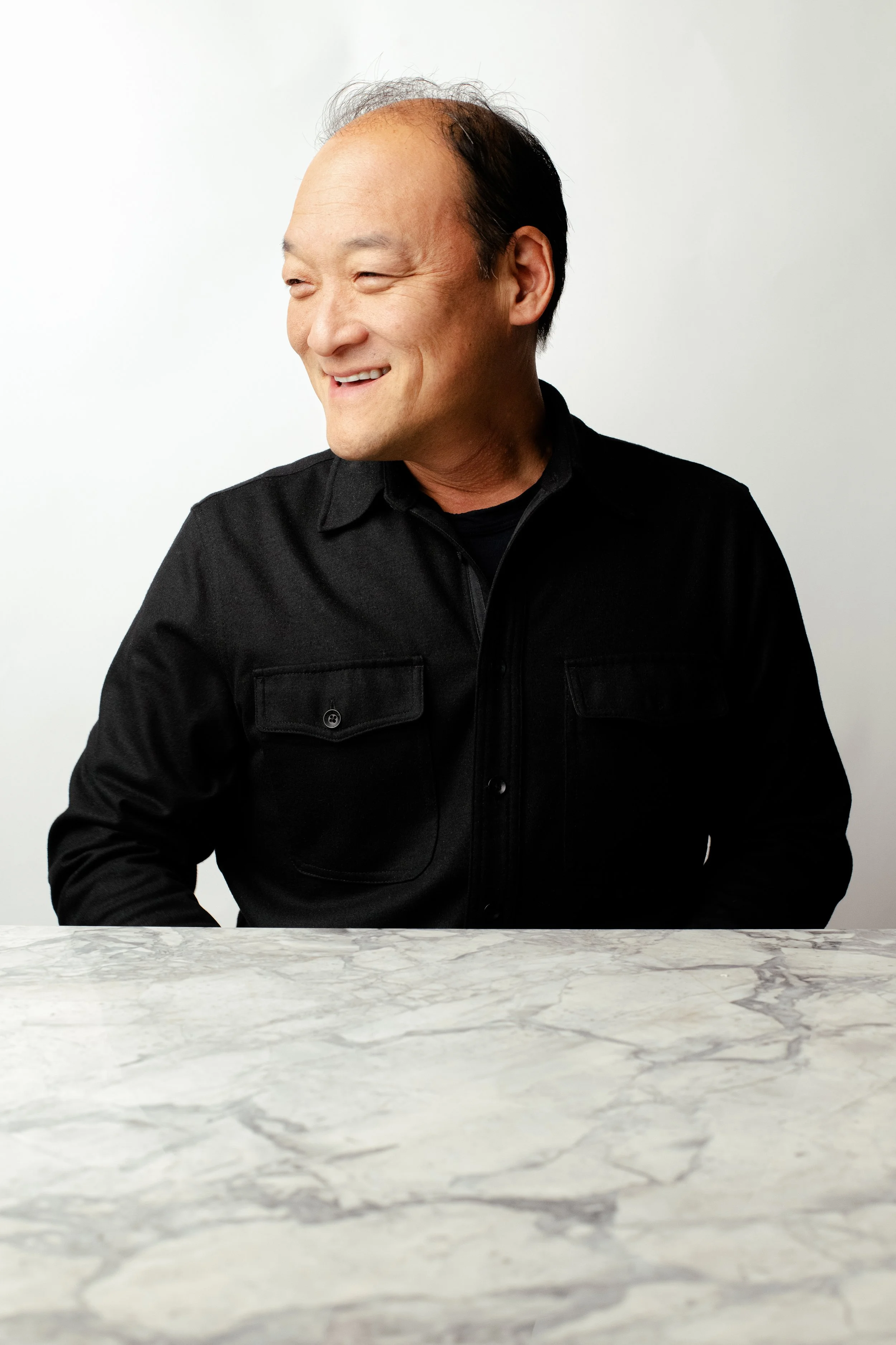 A smiling man with short dark hair, wearing a black jacket, sitting at a marble table against a white background