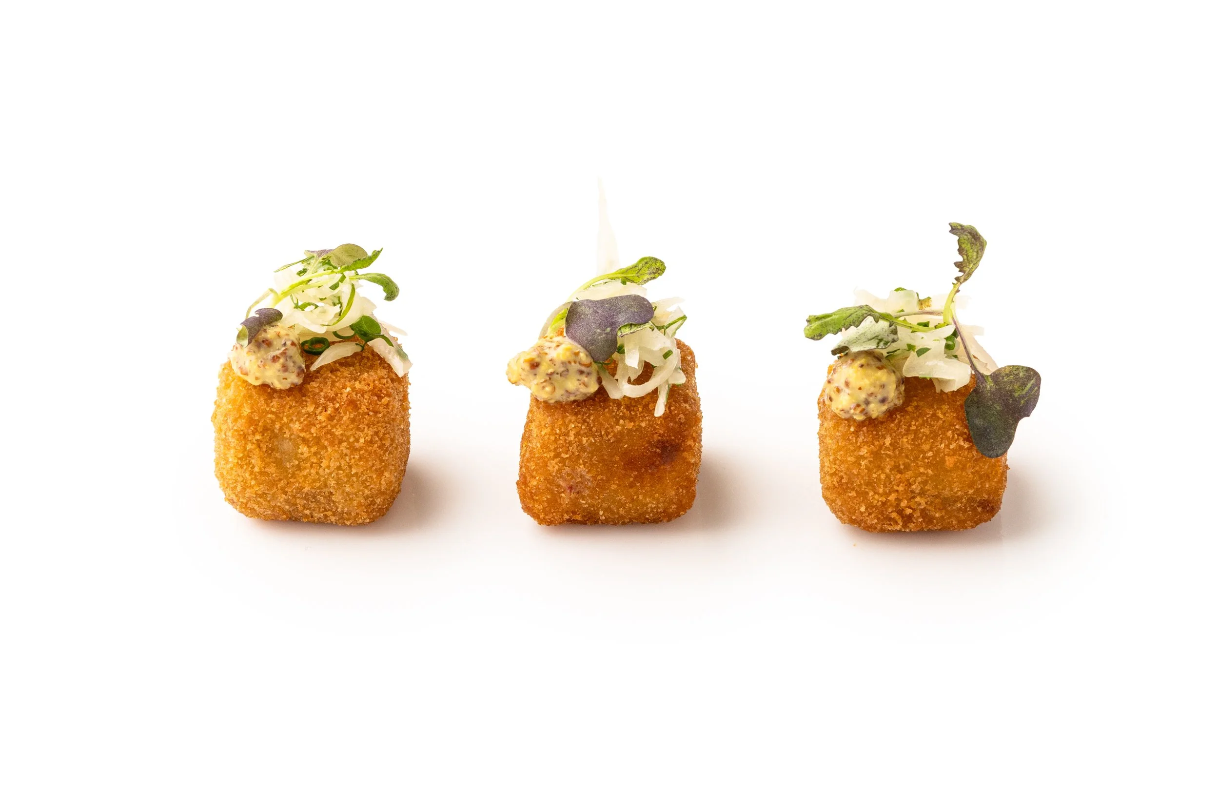 Three fried croquettes topped with microgreens and a dollop of mustard-based sauce on a white background.