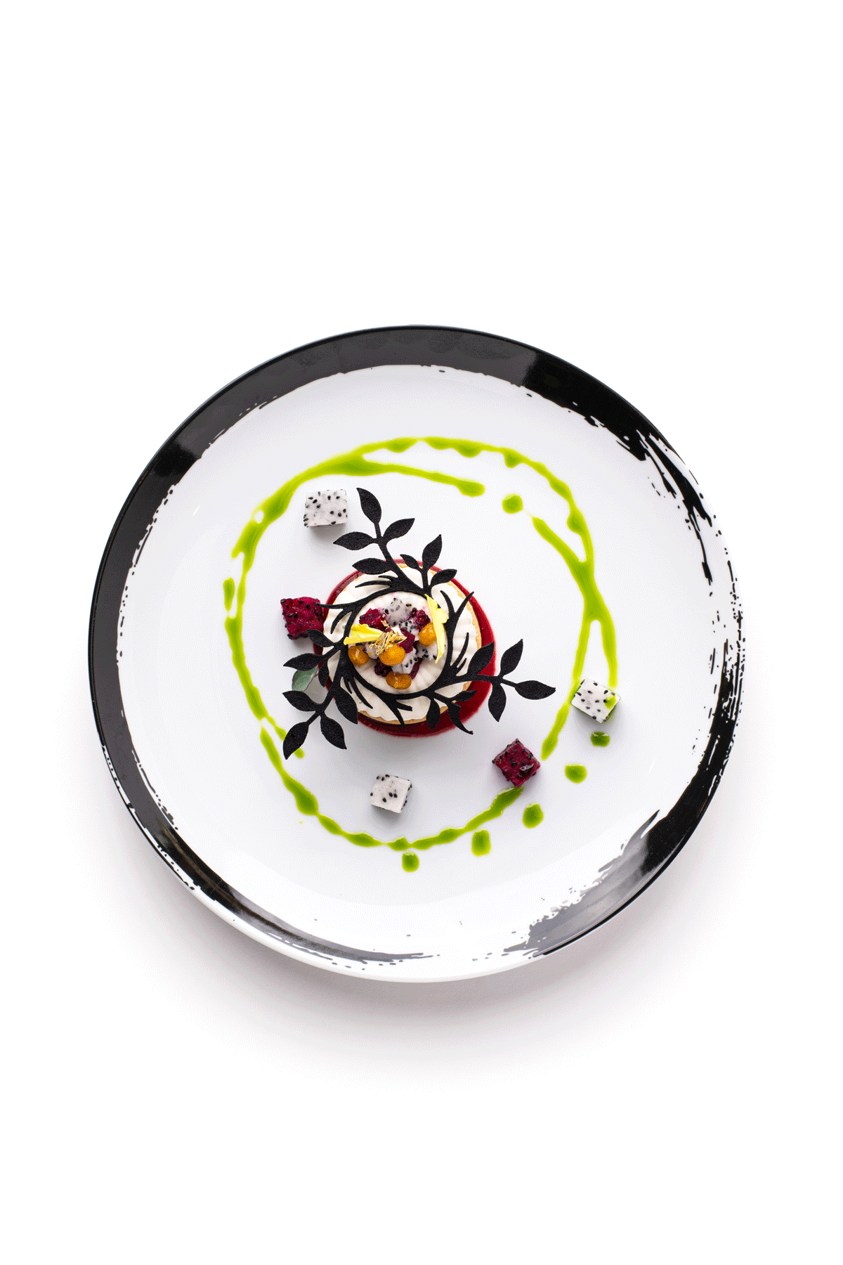 Elegant dessert plated with artistic decoration, including drizzles of green sauce, edible black leaf-shaped garnish, small cubes of red and white ingredients, and a central cream or mousse topped with berries and edible flowers on a white plate with