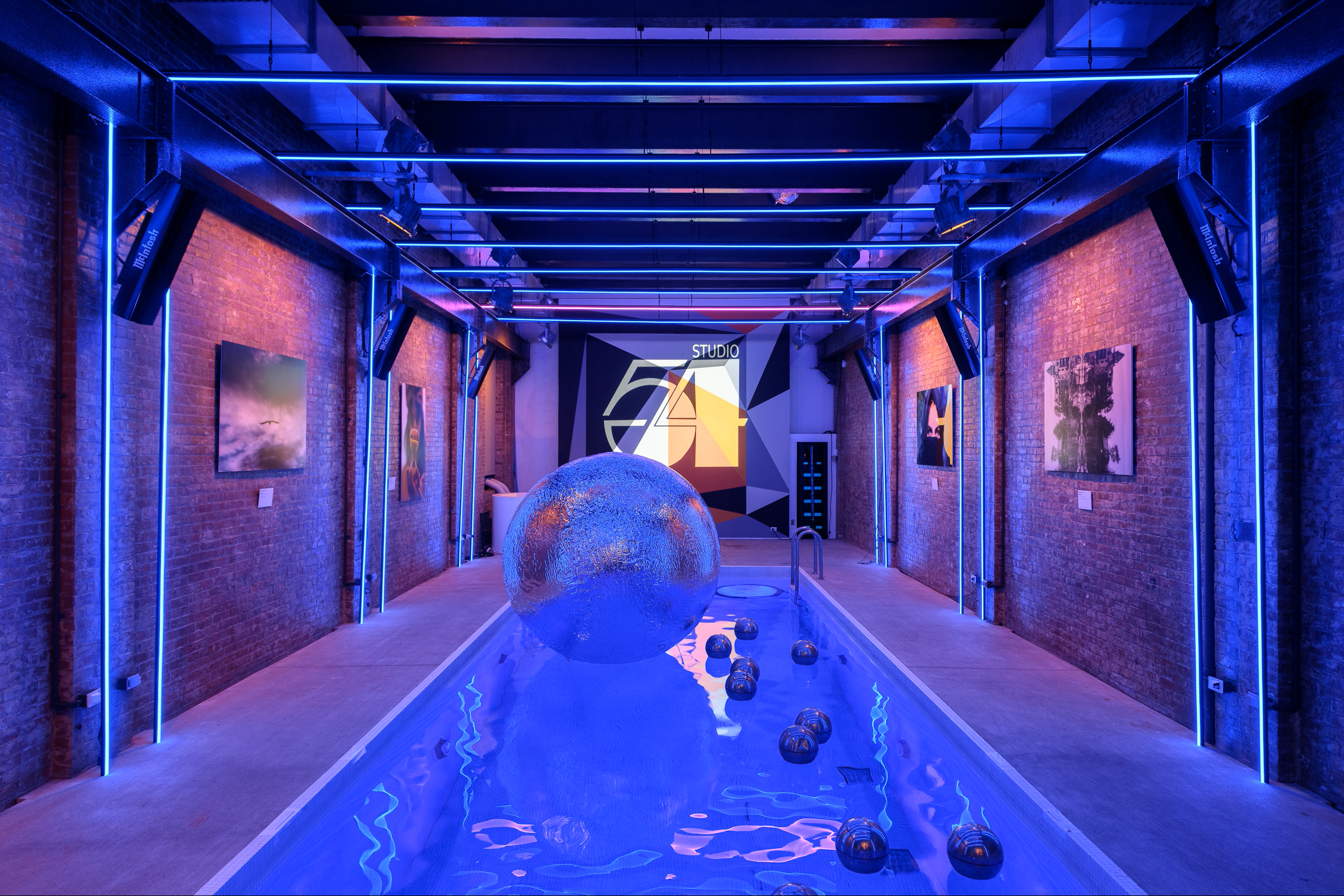 An art gallery with exposed brick walls illuminated by neon blue lighting, featuring several paintings on the walls and a water installation in the center with a large metallic sphere and smaller metallic spheres, leading to a digital display at the 