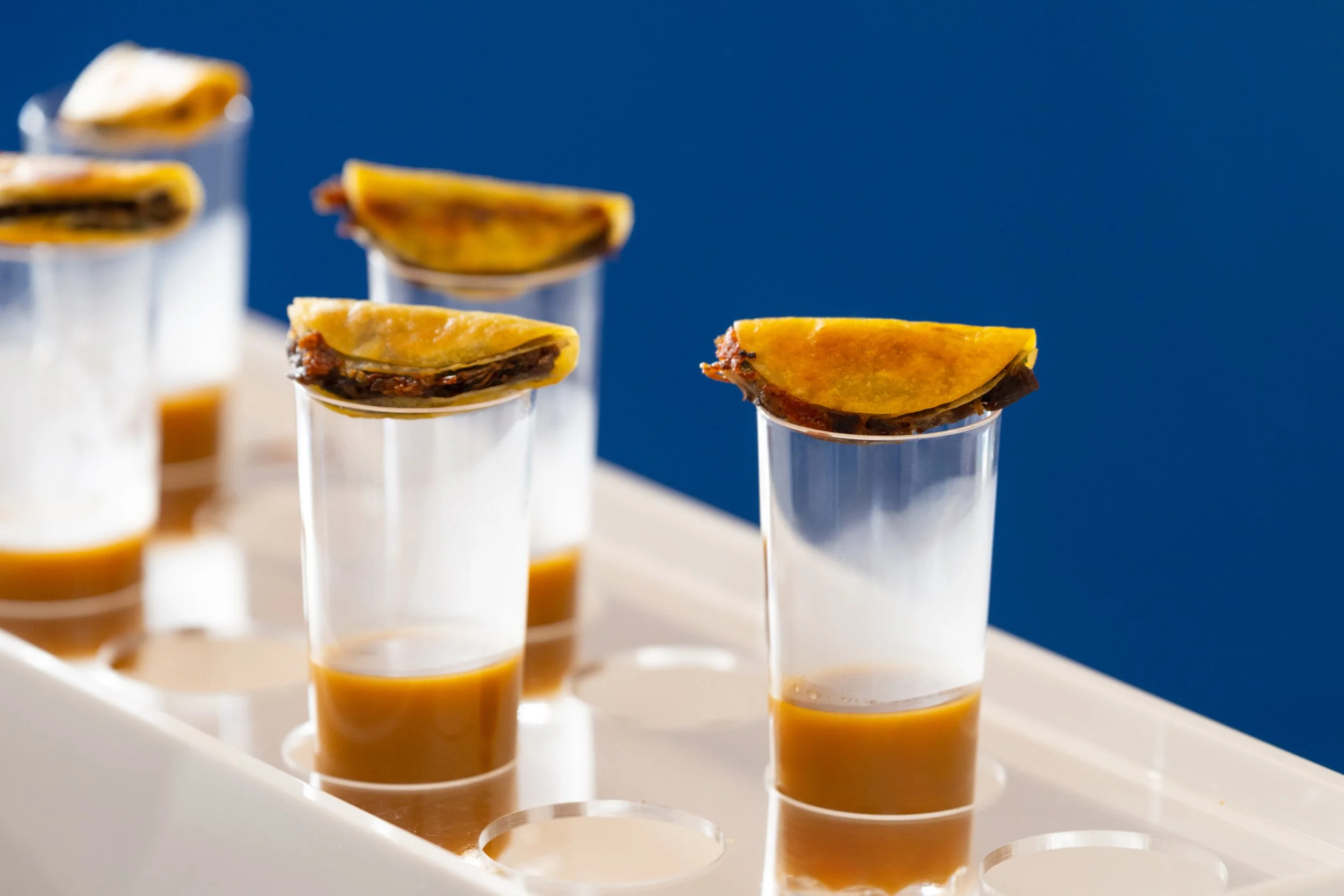 Small shot glasses filled with a brown liquid, each topped with a grilled lemon wedge, placed in a white holder against a blue background.