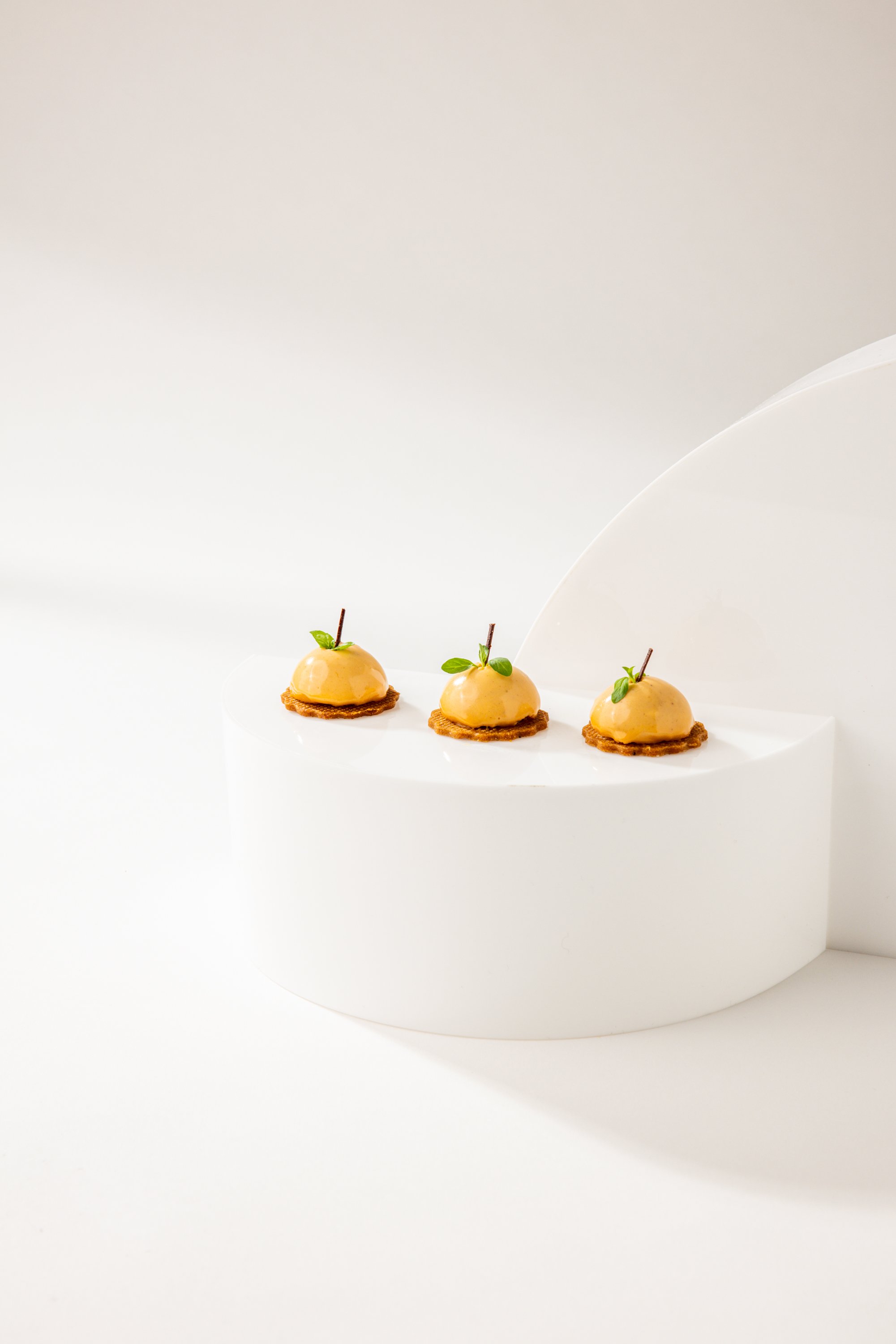 Three small, round desserts with a glossy yellow glaze, garnished with a tiny green leaf and a thin stick, placed on a white, curved surface.