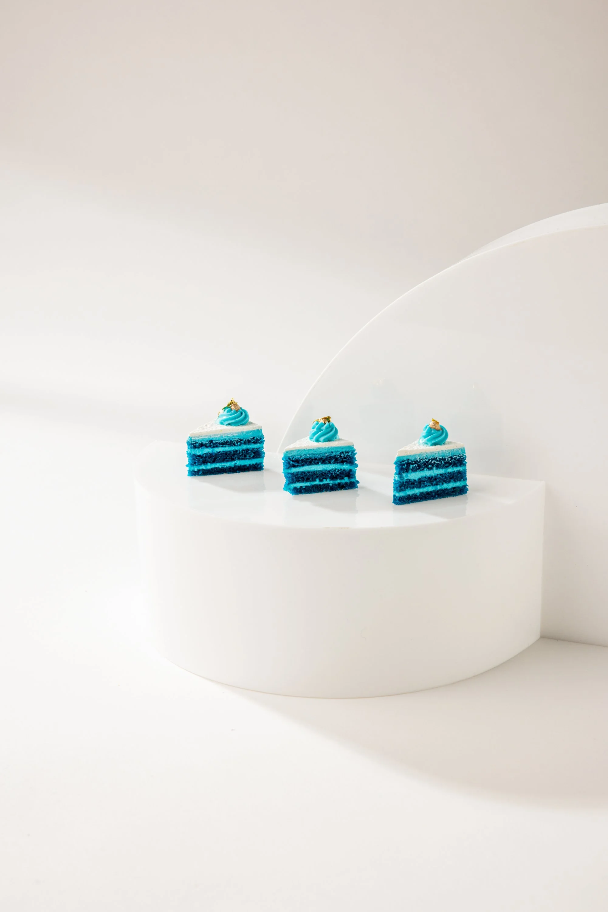 Three slices of blue layer cake with white frosting and blue swirl decoration on top, placed on a white curved display surface.