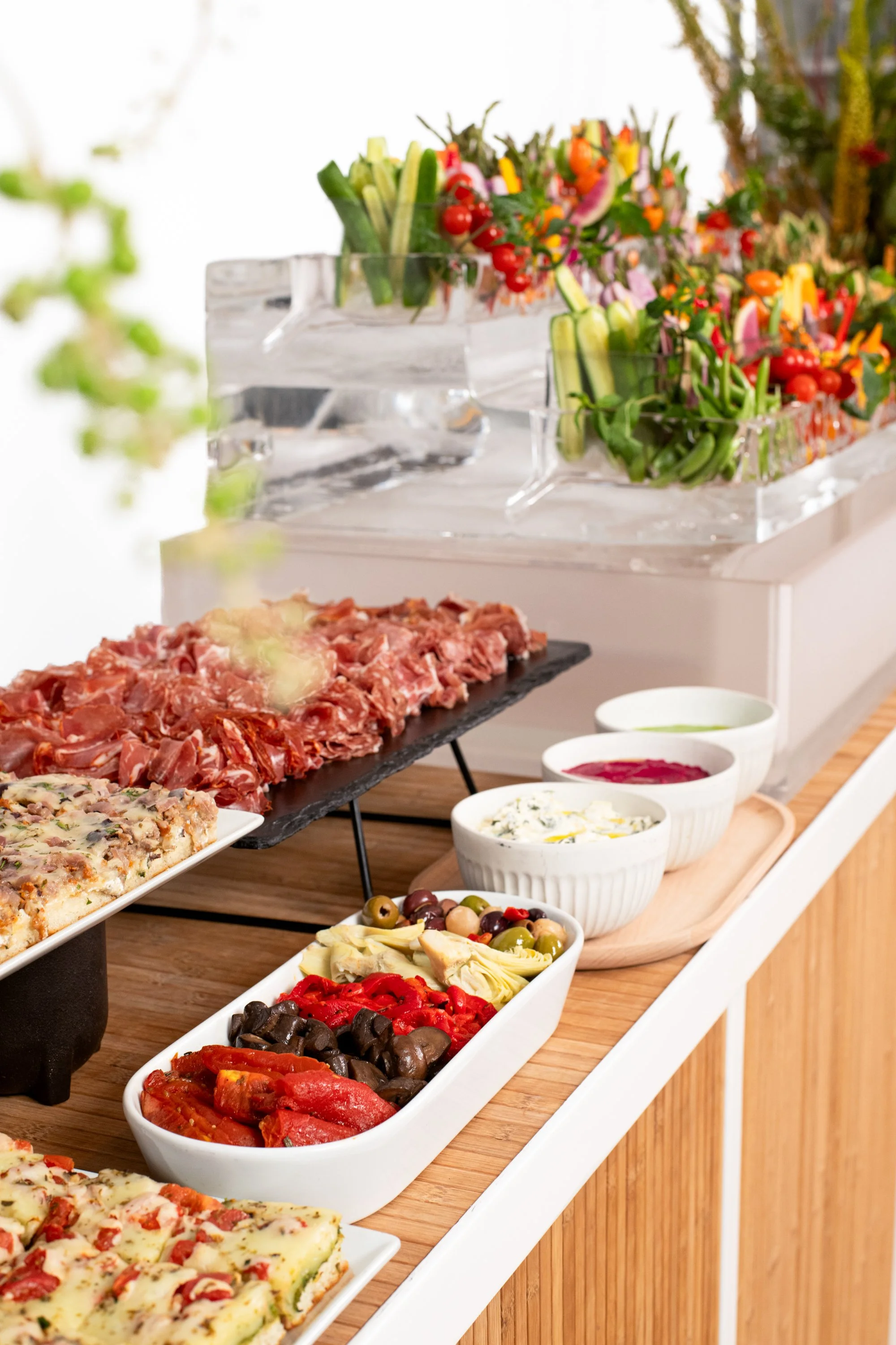 A buffet table with a variety of foods including sliced meats, cheeses, olives, pizza, and salads, with colorful vegetable arrangements in the background.