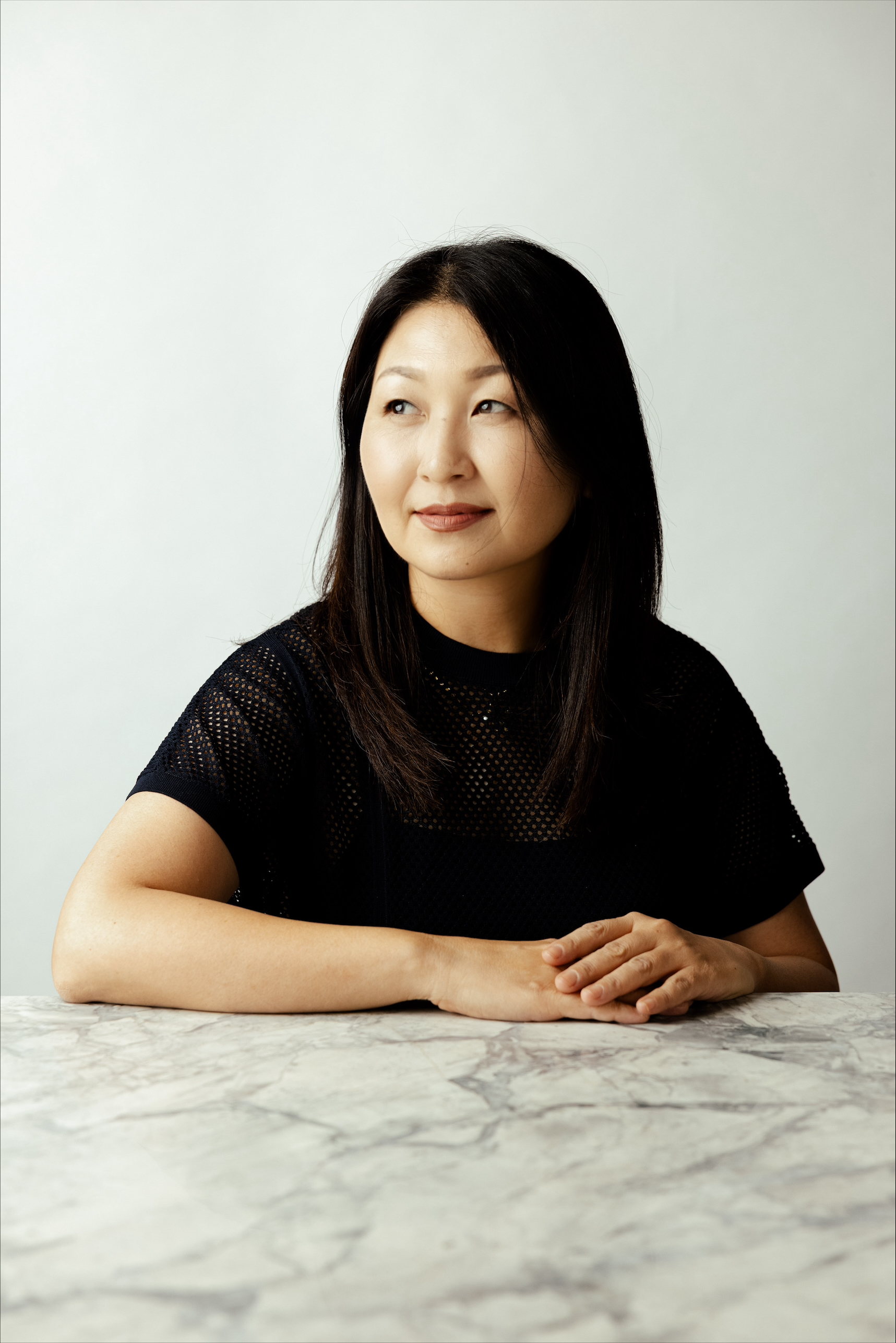 A woman with straight black hair wearing a black mesh top, sitting at a marble table with hands folded, looking to the side against a plain light grey background.