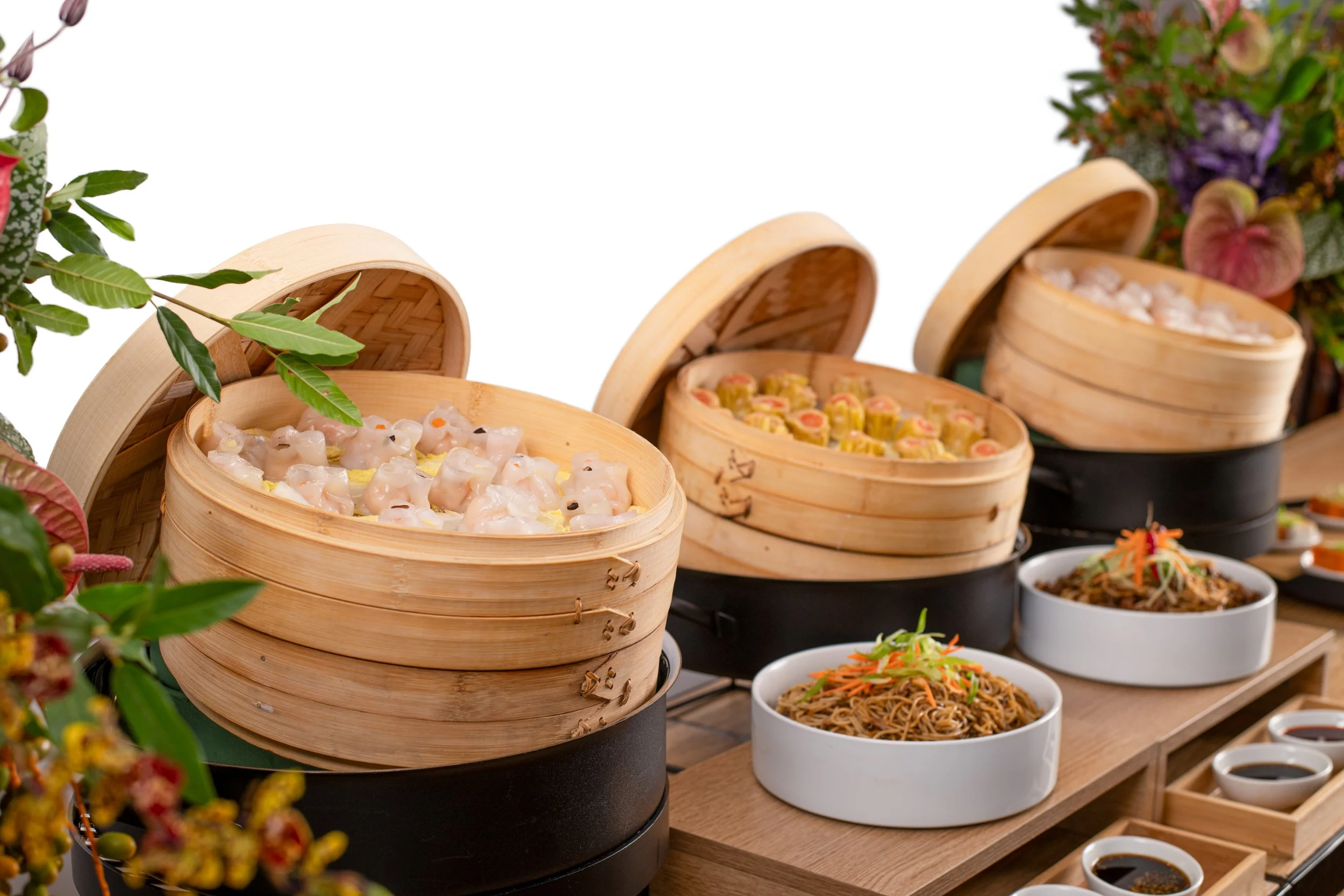 Assorted Asian dishes in bamboo steamers and bowls, including dumplings, noodles, and dipping sauces, on a wooden table.