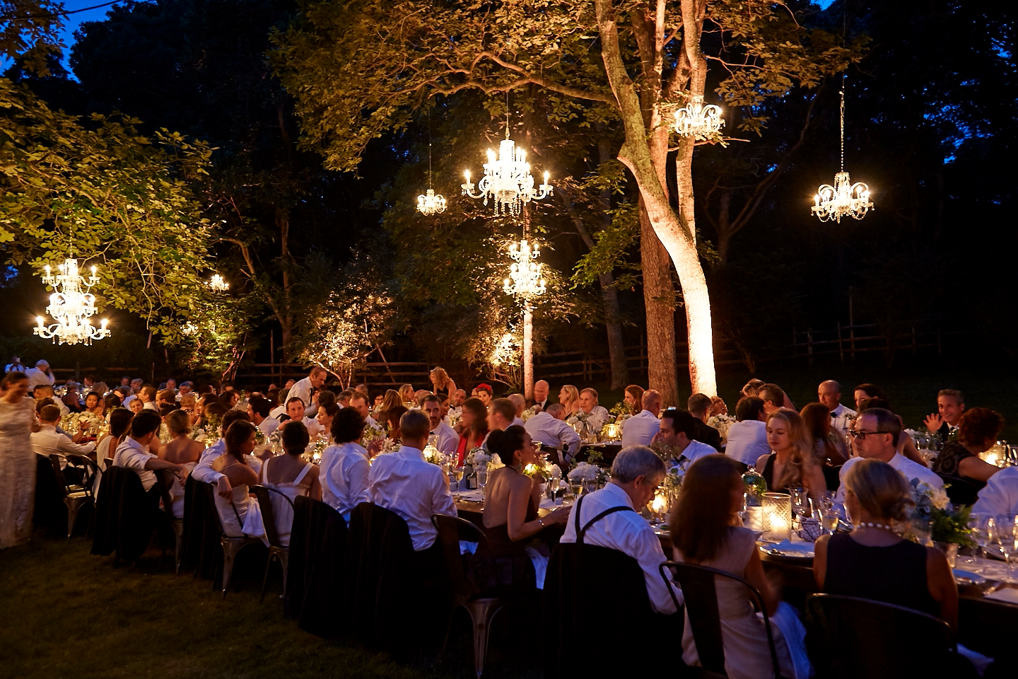 An outdoor evening dinner event with many guests seated at long tables, illuminated by chandeliers hanging from trees.