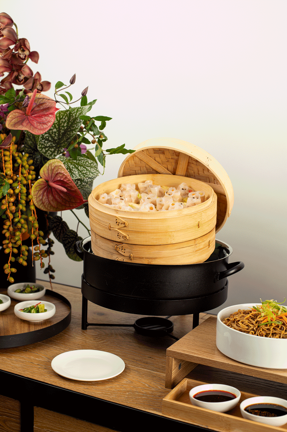 A bamboo steamer with dim sum on an electric hot plate, surrounded by dishes of sauces, noodles, and a plant with colorful leaves.