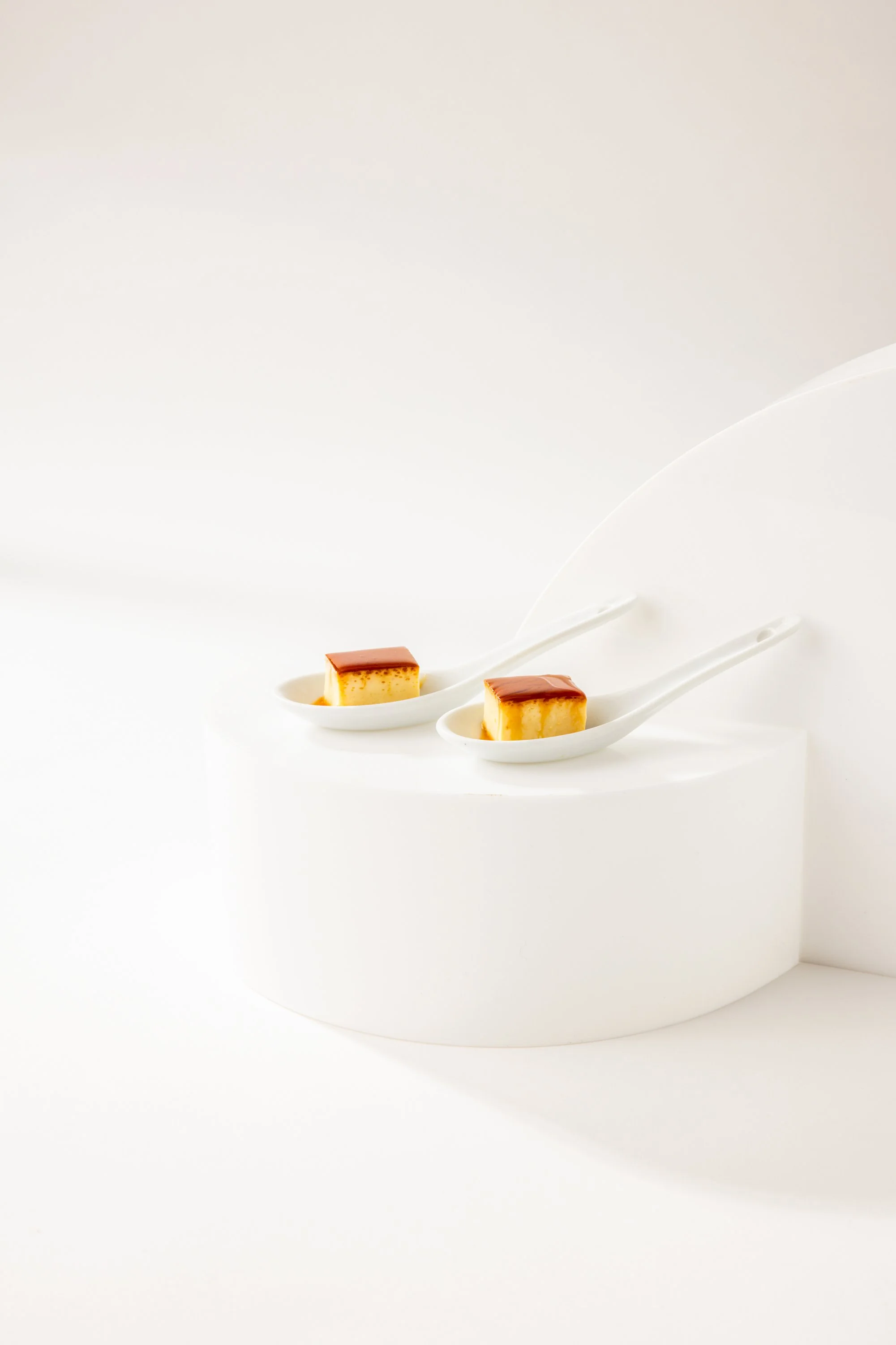 Two slices of flan caramel dessert in white ceramic spoons on a white display surface.
