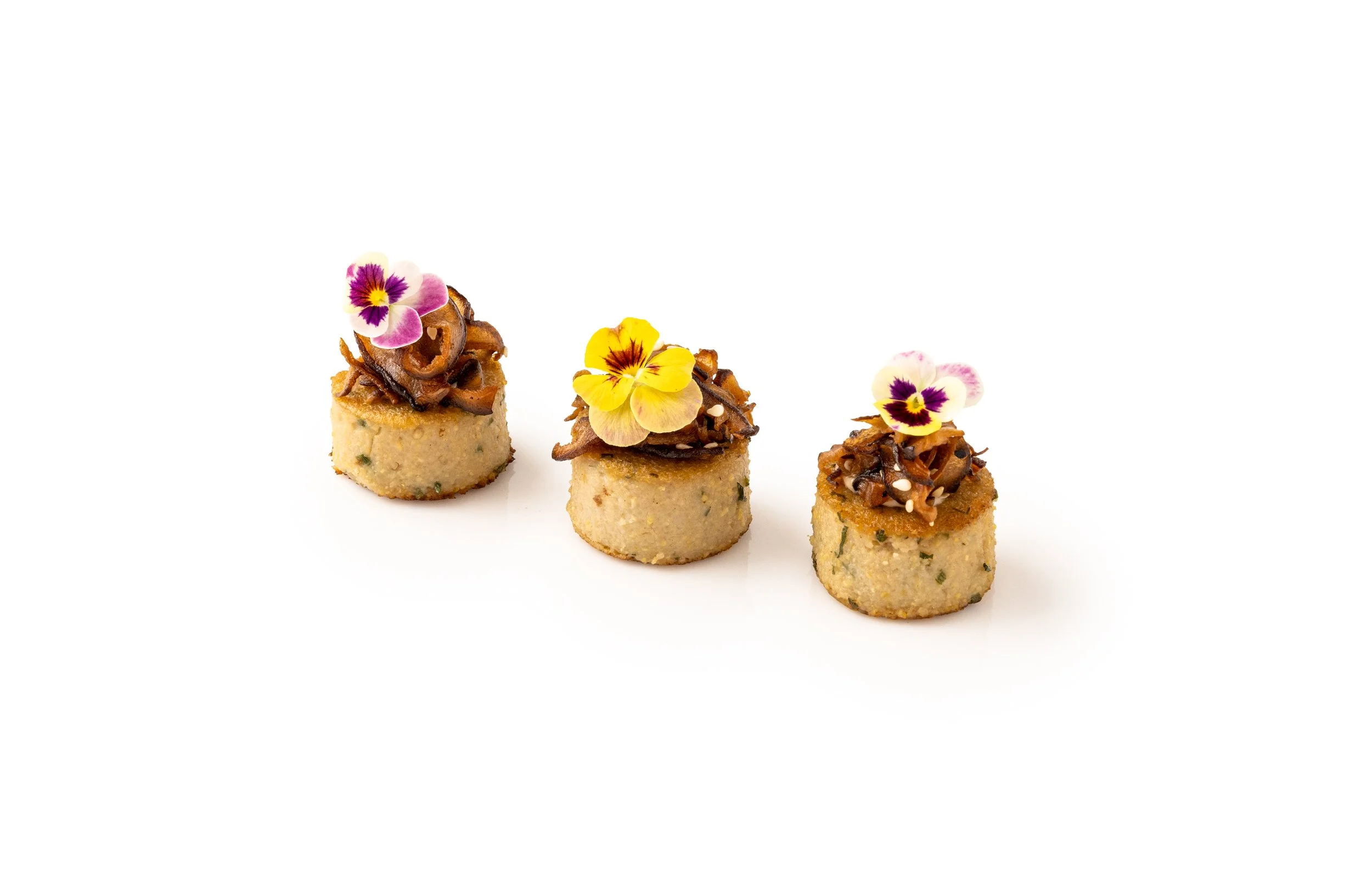Three gourmet mini savory cakes topped with caramelized onions and edible flowers, arranged in a row on a white background.