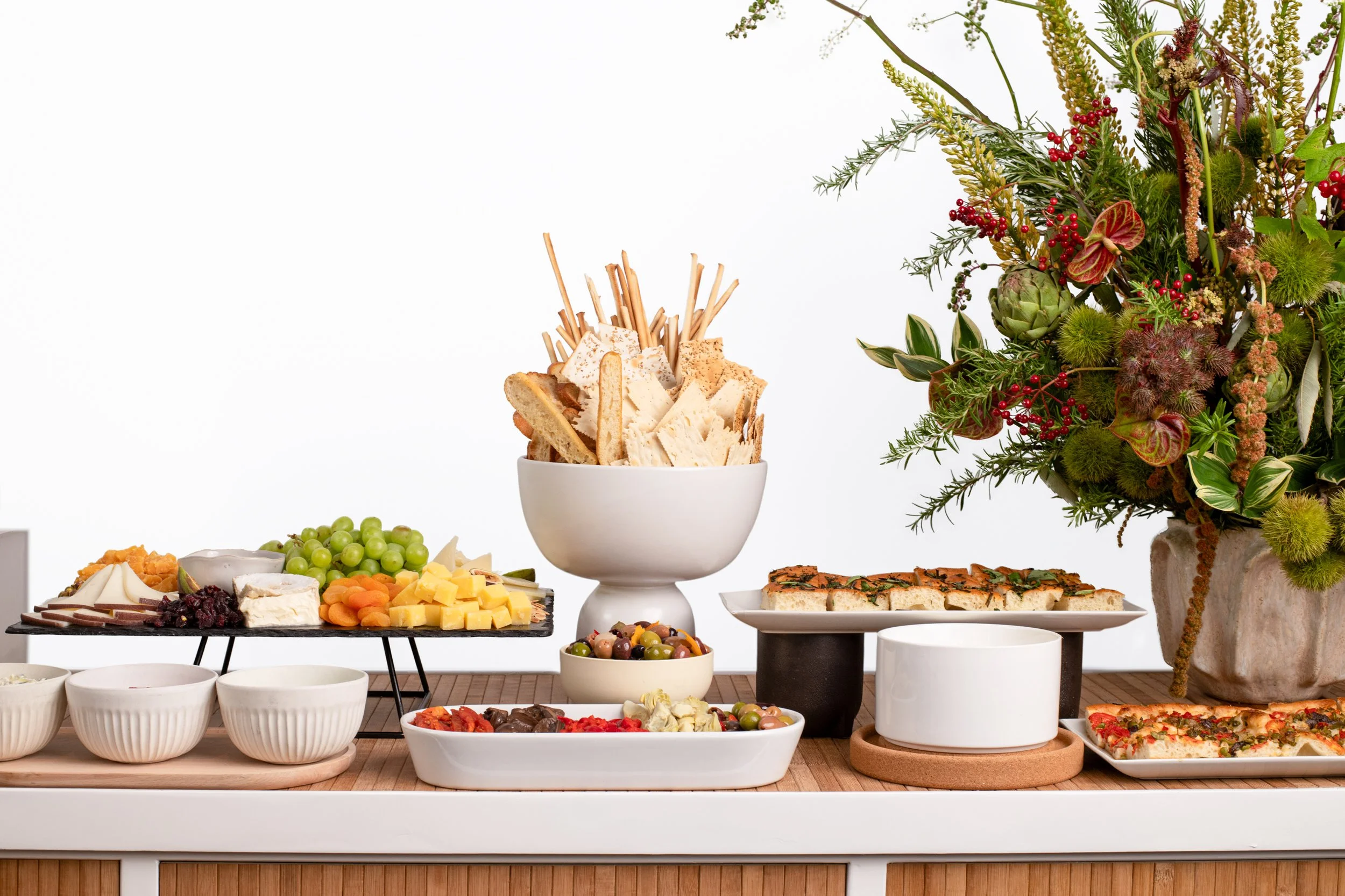 A variety of snacks and finger foods on a table, including cheese, crackers, grapes, olives, vegetables, pizza, and an appetizer platter, with a large floral arrangement on the right.