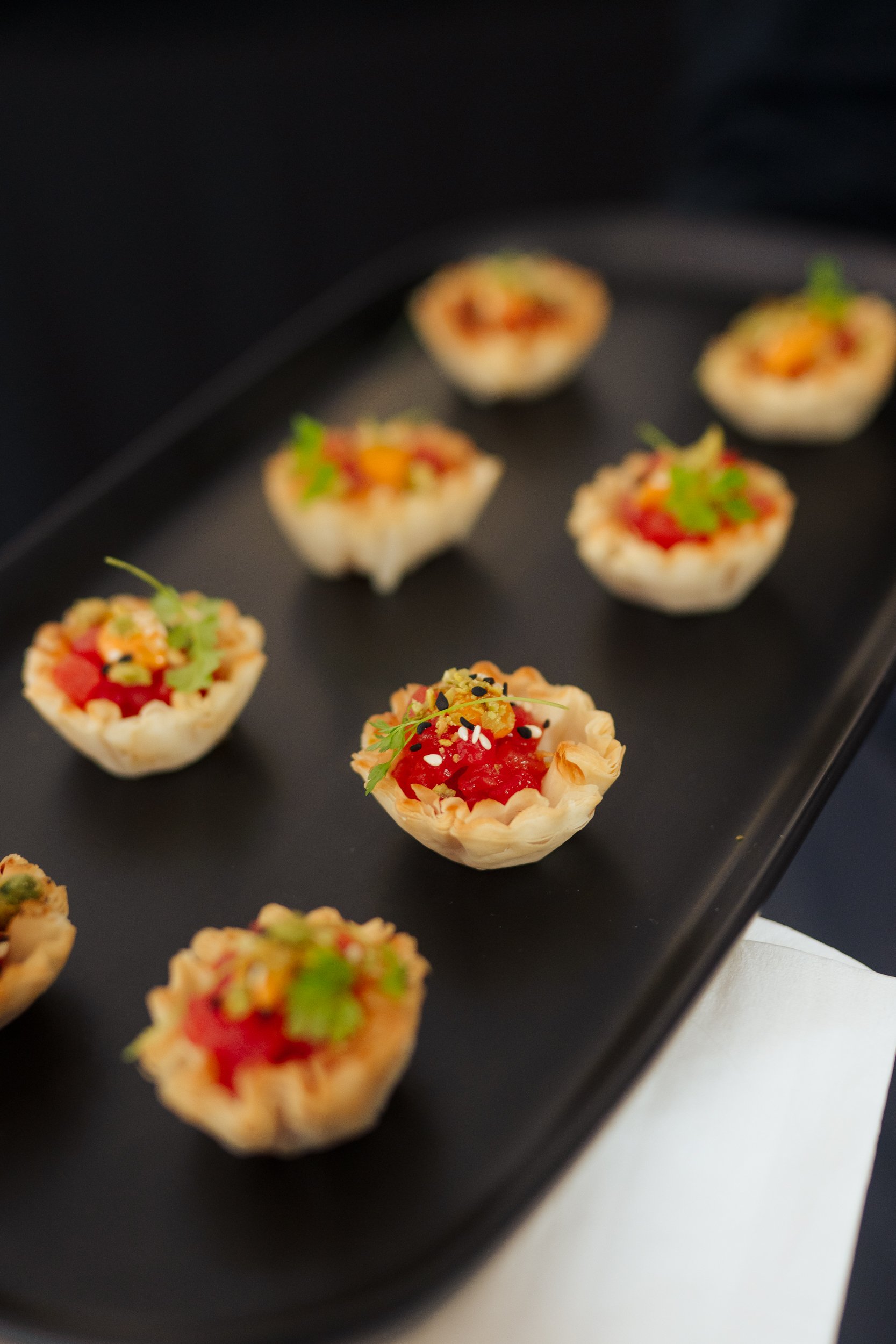 Small savory tartlets with various toppings, garnished with herbs, displayed on a black serving tray.