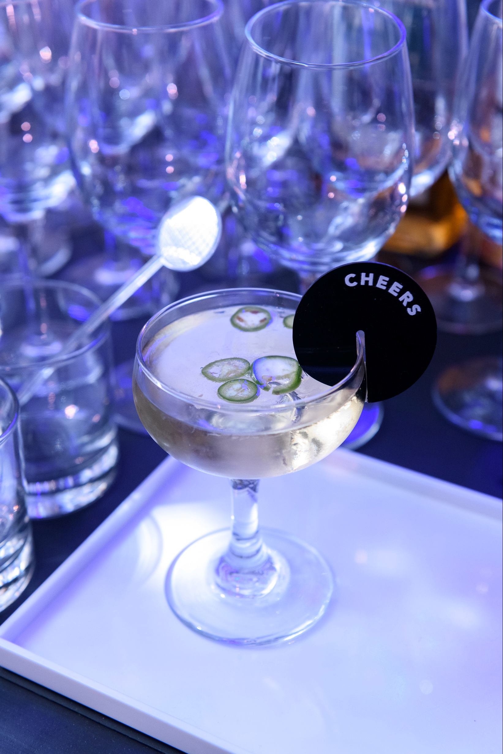 A cocktail with slices of cucumber garnished with a black 'Cheers' sign, set on a white tray surrounded by empty glasses.