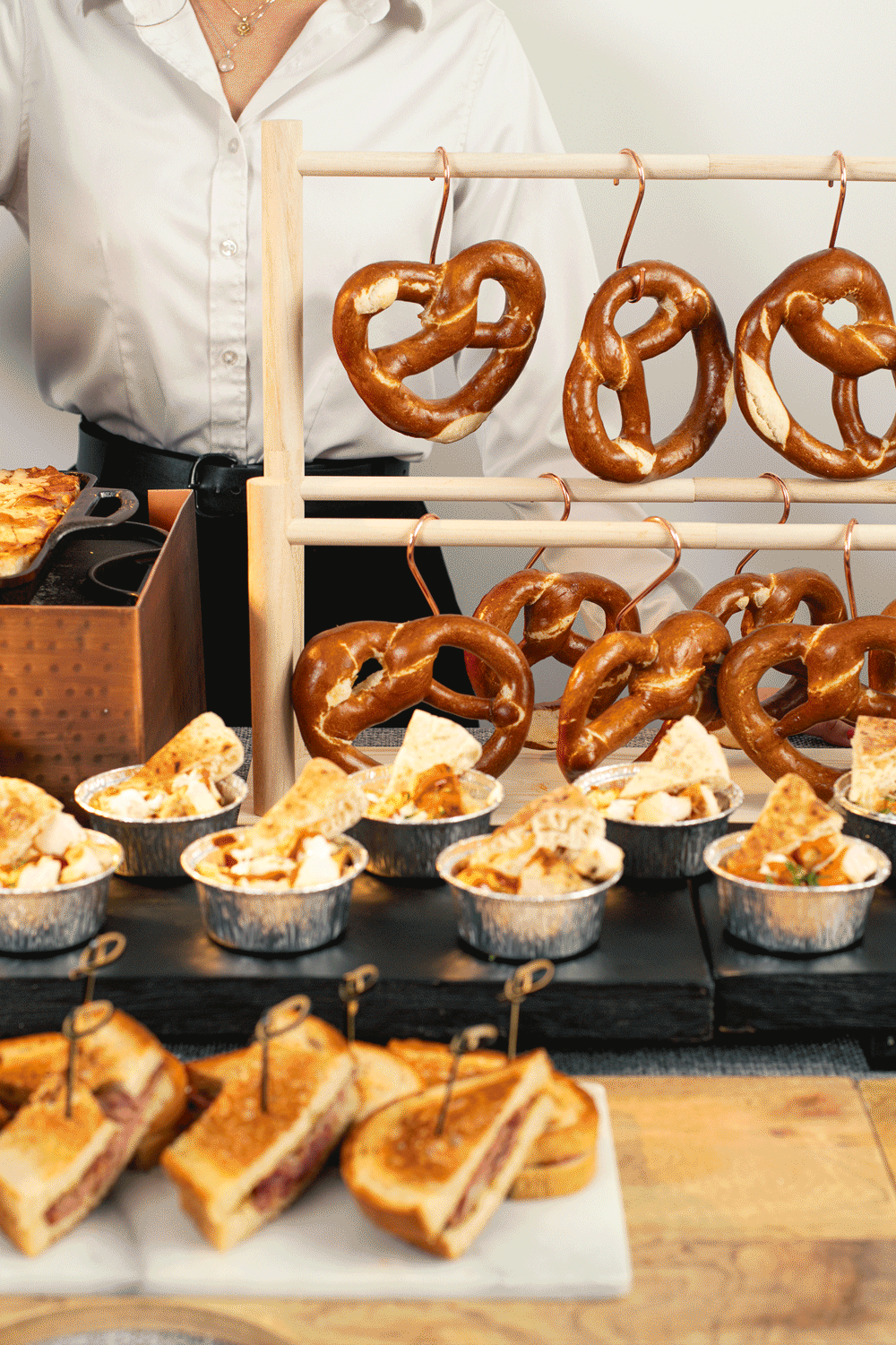 Pretzels hanging on a stand at a food market, with sandwiches and small bowls of food in the foreground.