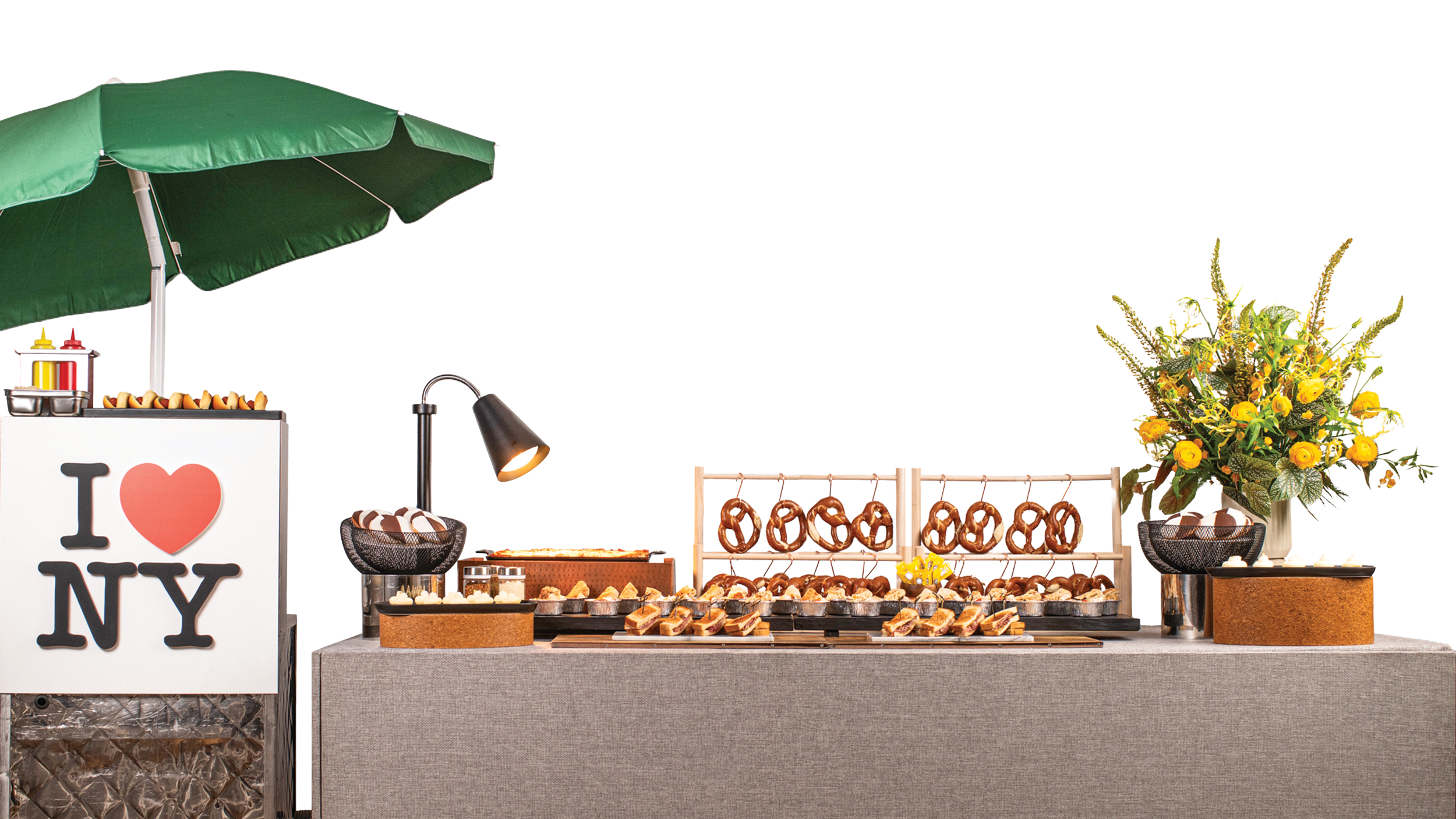 Food and drink setup at an outdoor event with a green umbrella, a sign reading 'I love NY', a lamp, a flower arrangement, and various snacks including pretzels and sandwiches on a table.