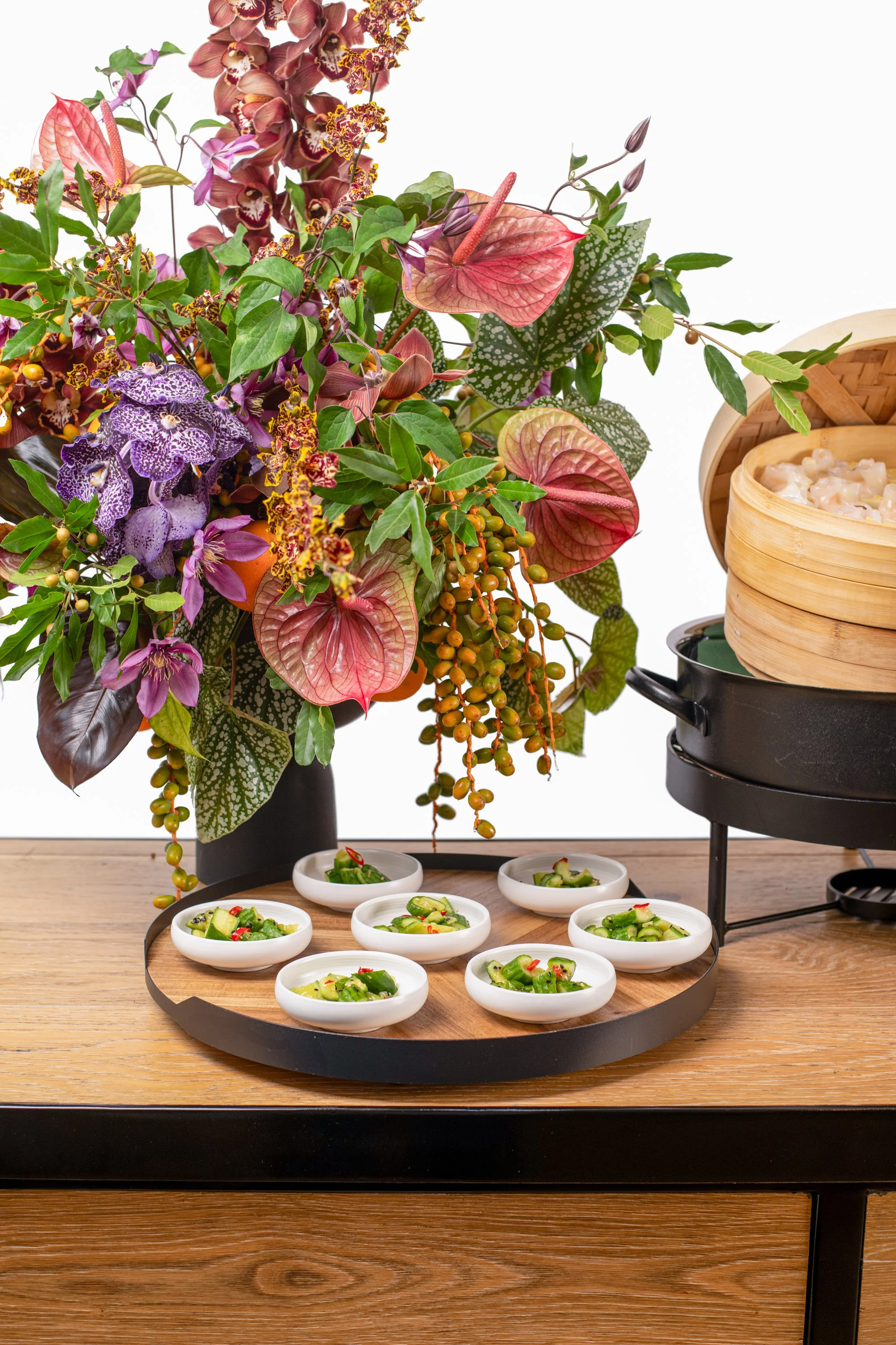 A large floral arrangement with pink, purple, and green flowers and leaves on a table, with small bowls of sliced cucumber with red chili, and a bamboo steamer and black pot in the background.