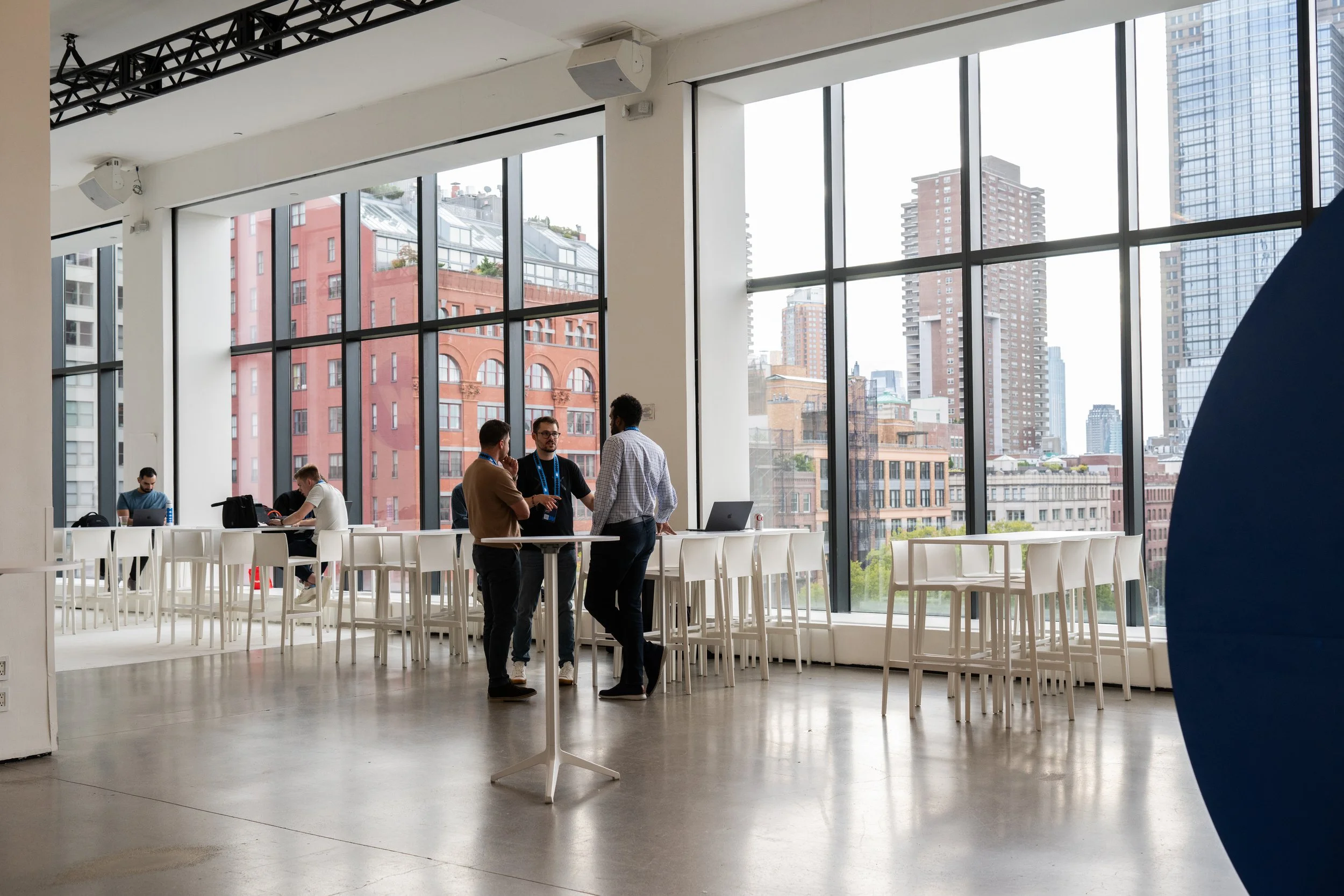 Three people are standing and talking near a high table in an office space with large windows showing a cityscape of tall buildings. Two people are sitting at a table, working on laptops, while another person is partially visible on the right side of